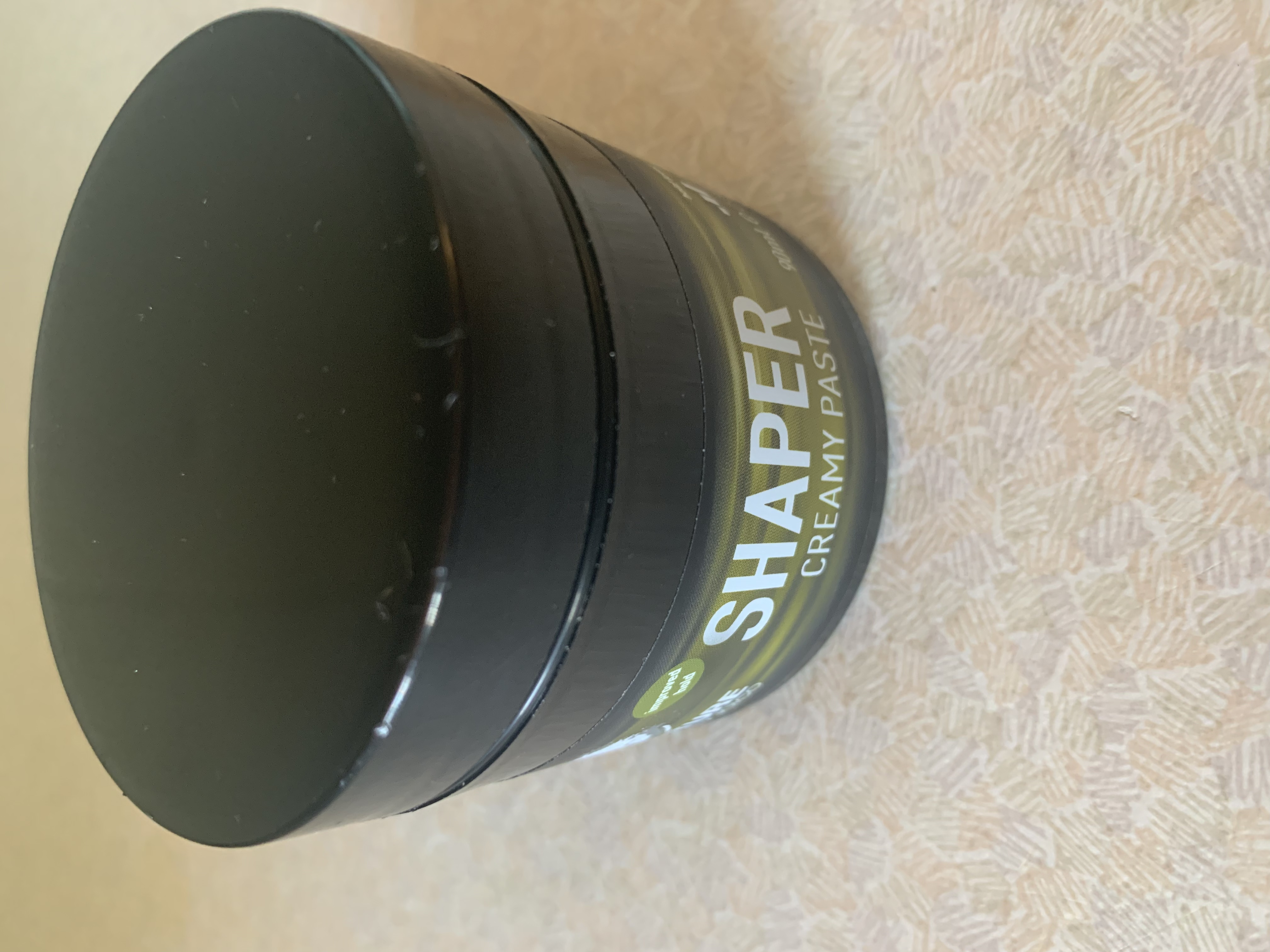A short cylindrical black plastic tub with a Green label that has Structure by Joico Shaper written in dark blue writing on it, on a white background.