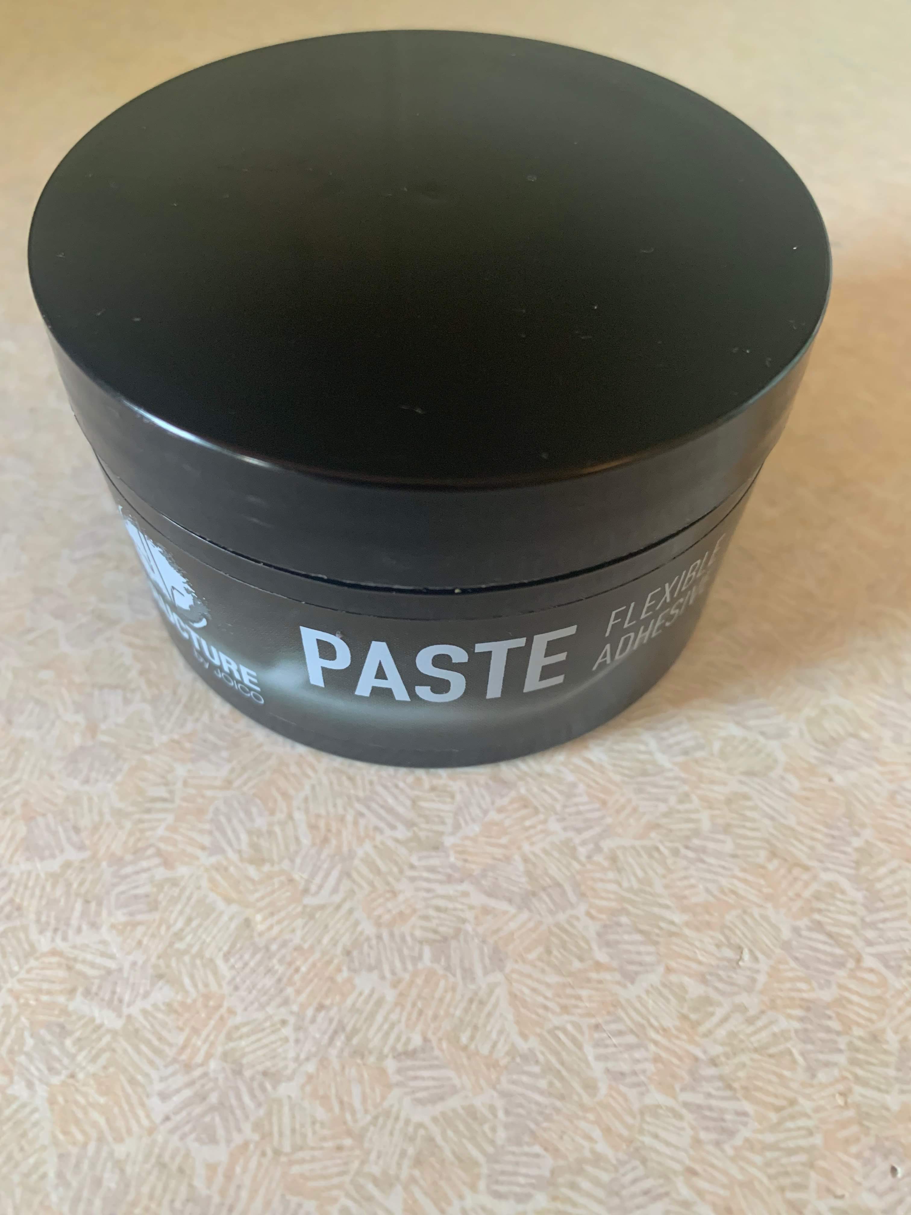A short cylindrical black plastic tub with a Grey label that has Structure by Joico Paste written in dark blue writing on it, on a white background.