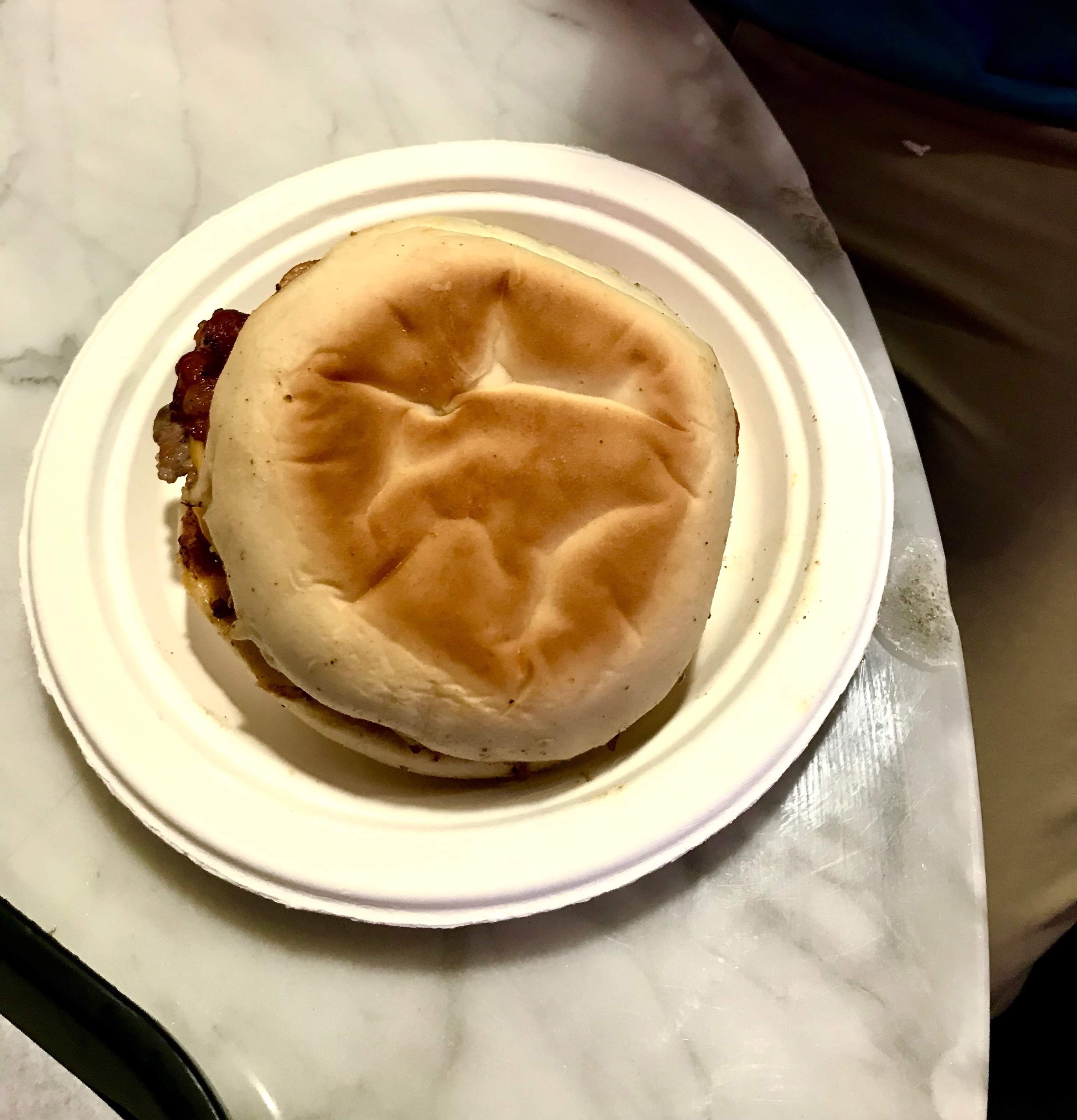 A large circular golden brown English Muffin full of some bright white pieces of Egg on a small circular white paper plate on a light wooden table, on a white background.