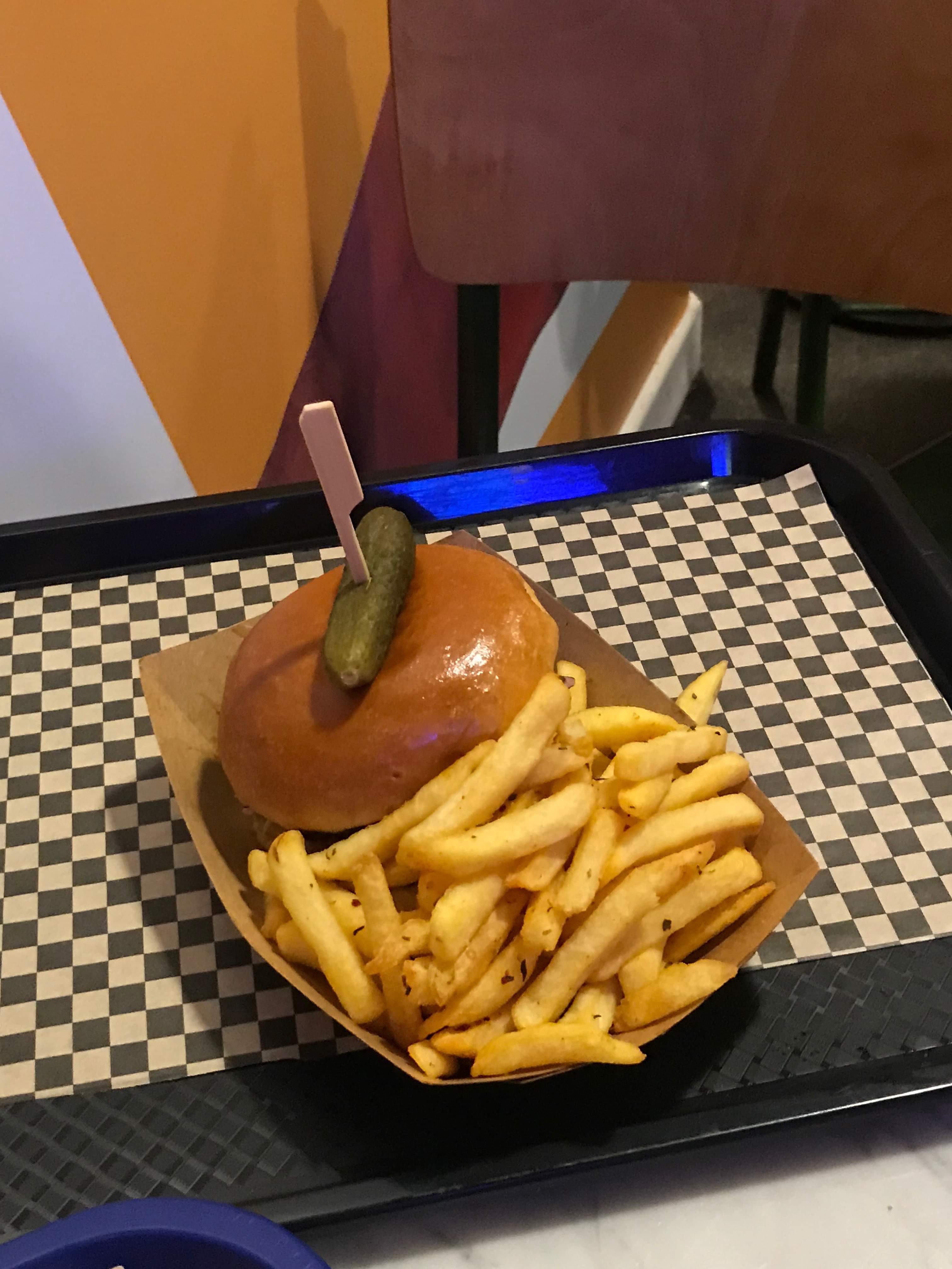 A large circular golden brown Bread Roll full of some dark brown pieces of Beef covered in some dark brown sauce next to a pile of golden brown seasoned fries on a small circular white paper plate on a light wooden table, on a white background.