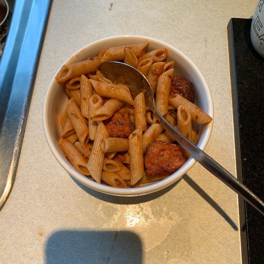 Some golden brown tubes of pasta and some pieces of dark brown sausage covered in some dark red sauce inside a white ceramic bowl on a white worktop, on a white background.