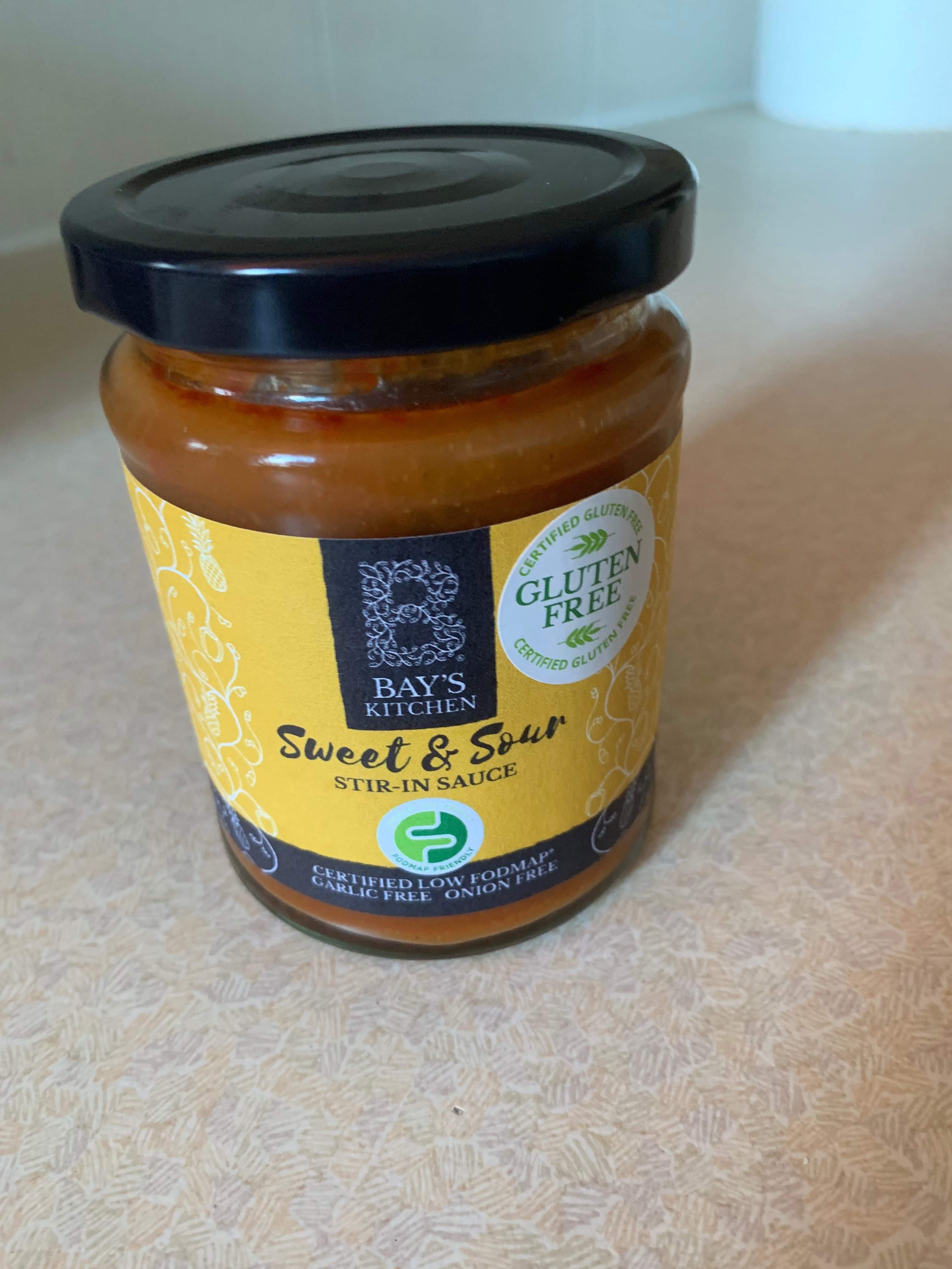 A short cylindrical glass jar full of some dark brown liquid with a black lid and a bright yellow label that has Bay’s Kitchen Sweet & Sour Stir-In Sauce written in bold black writing on it, on a white background.