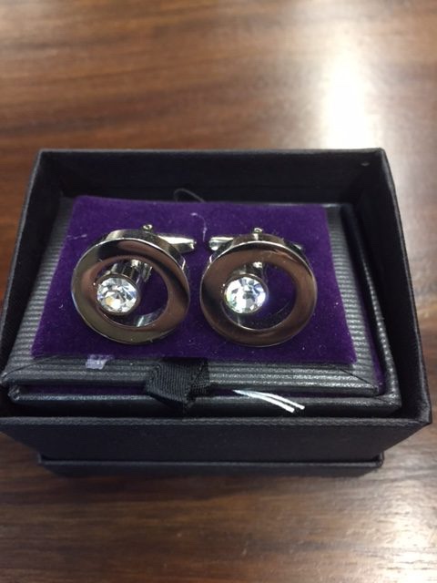 A short rectangular black gift box that has two circular shaped cufflinks with a diamond pattern on them inside it, on a white background.