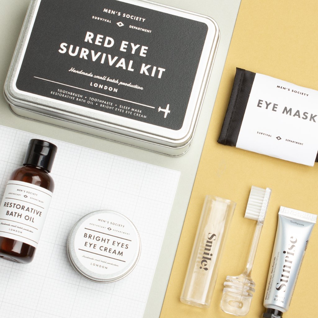 A tall cylindrical silver tin that has Men's Society Red Eye Survival Kit written in bold black writing on it next to a white Eye Mask and a short cylindrical glass bottle full of some dark coloured liquid, on a white background.