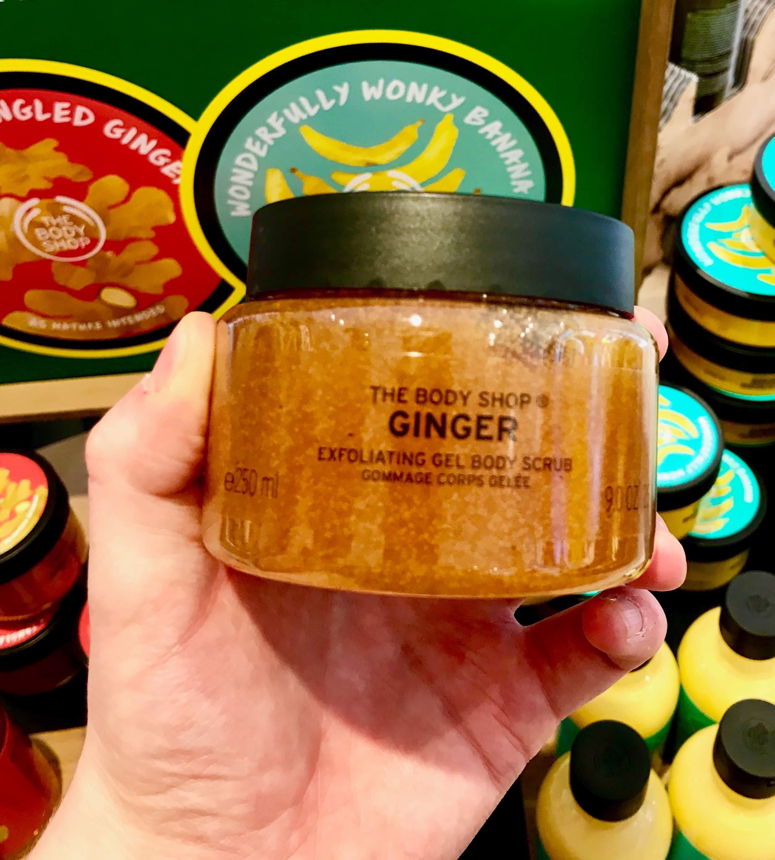 A short cylindrical clear plastic tub full of some bright orange liquid with a black lid that has The Body Shop Ginger Body Scrub written in bold black writing on it, on a white background.