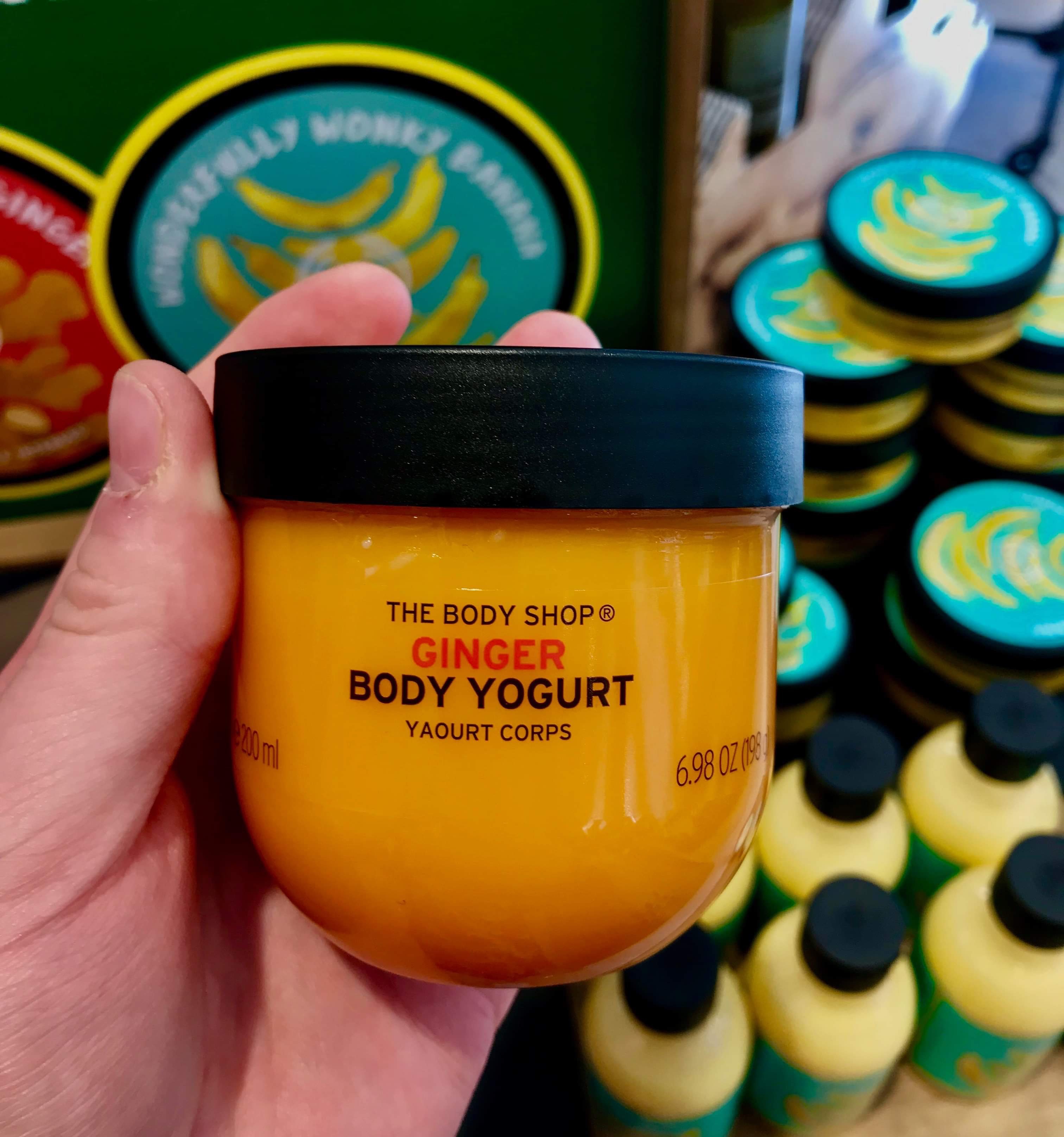 A short rounded rectangular clear plastic tub full of some bright orange liquid with a black lid that has The Body Shop Ginger Body Yoghurt written in bold black writing on it, on a white background.