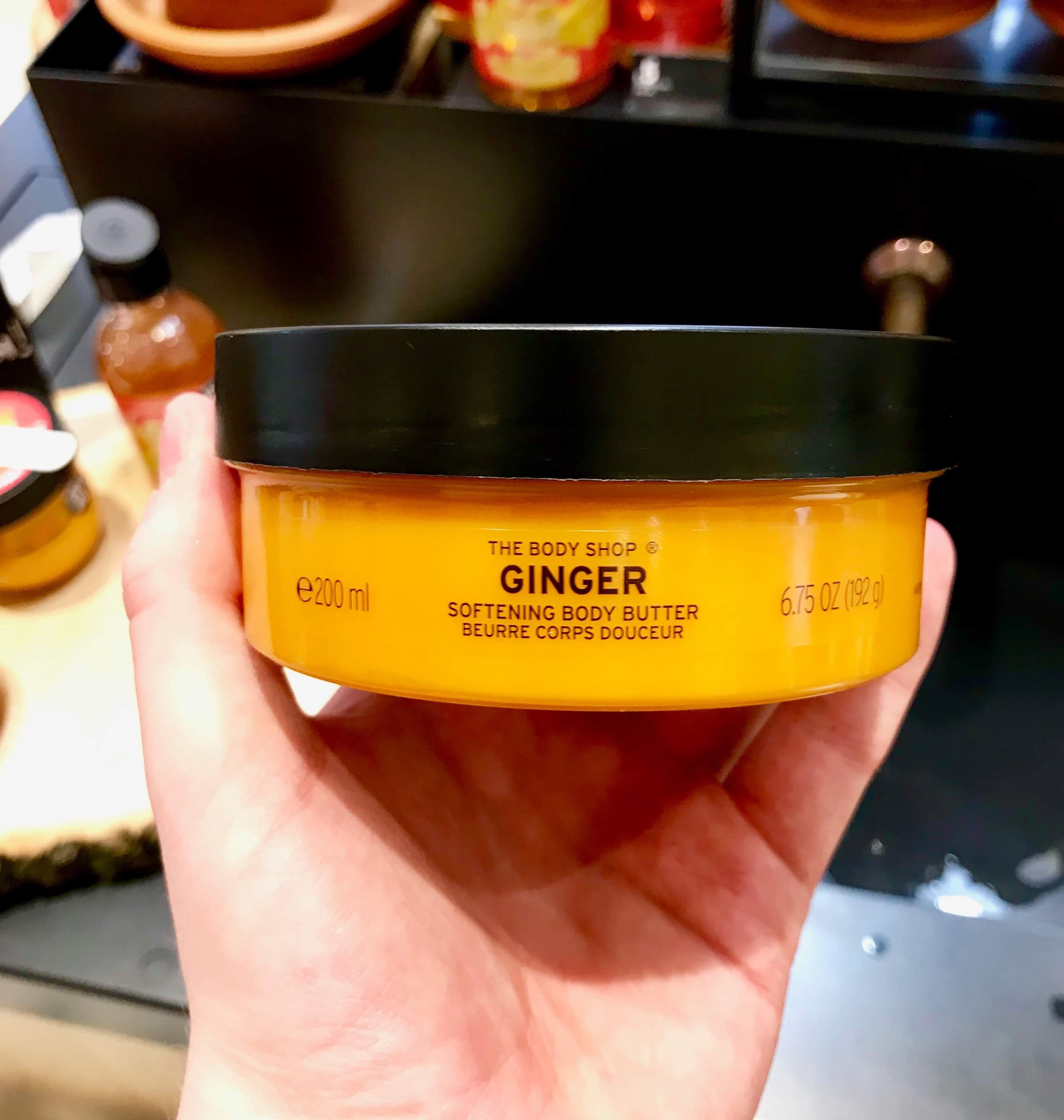 A short cylindrical clear plastic tub full of some bright orange liquid with a black lid that has The Body Shop Ginger Softening Body Butter written in bold black writing on it, on a white background.
