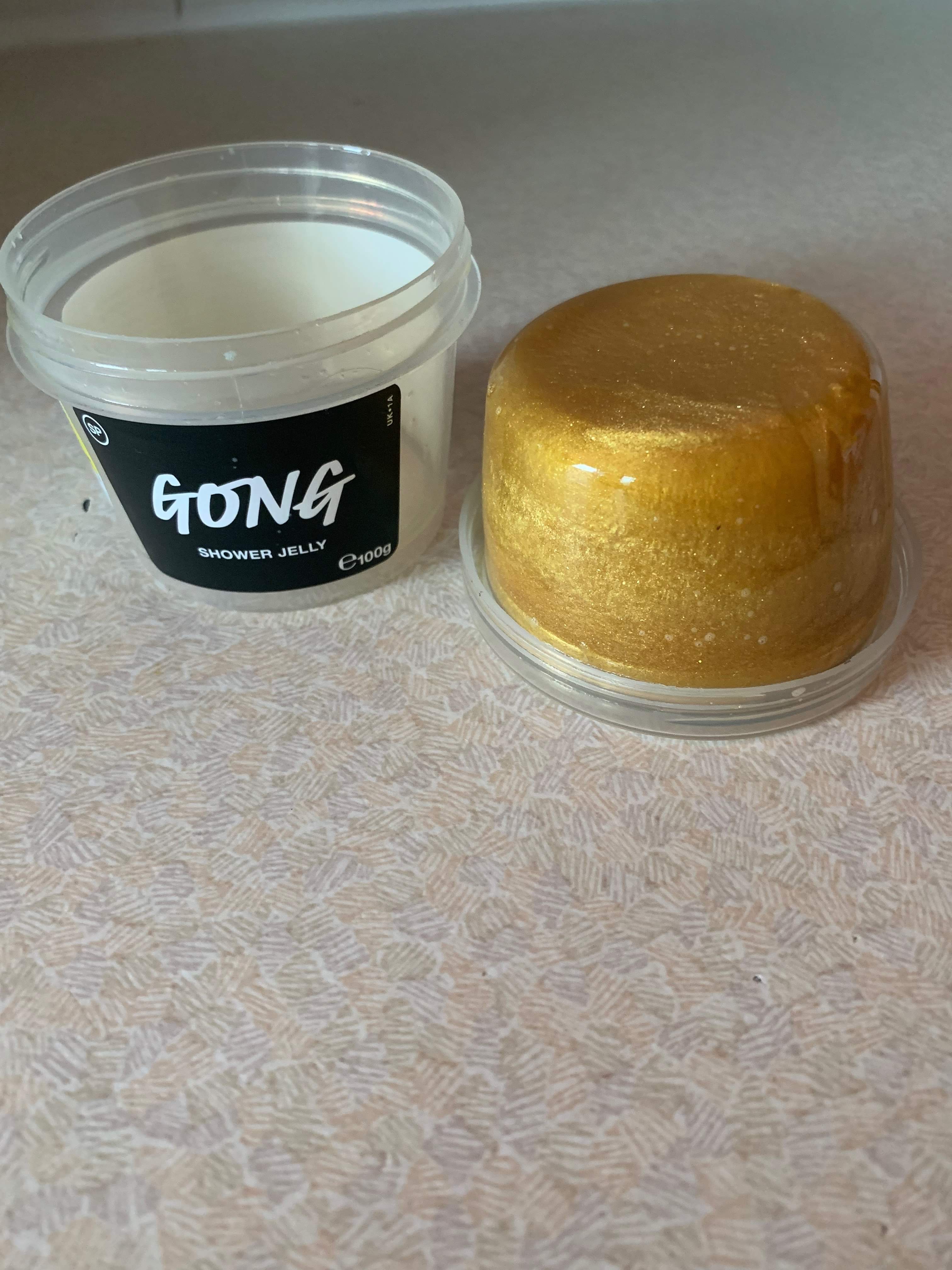 A short cylindrical block of shimmering gold shower jelly next to a short cylindrical clear plastic tub with a black label that has Lush Gong Shower Jelly written in bold white writing on it, on a white background.