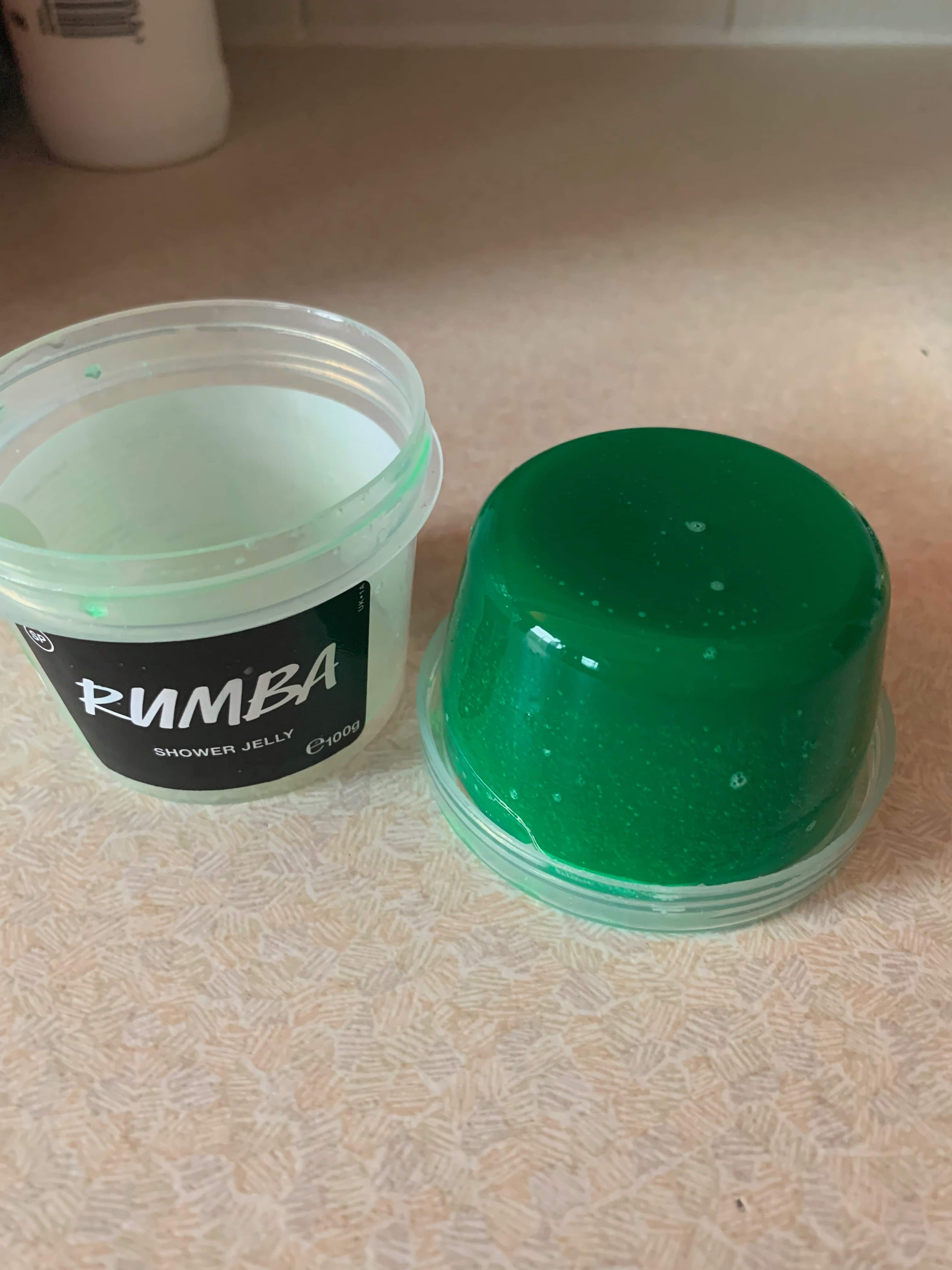 A short cylindrical block of dark green shower jelly next to a short cylindrical clear plastic tub with a black label that has Lush Rumba Shower Jelly written in bold white writing on it, on a white background.