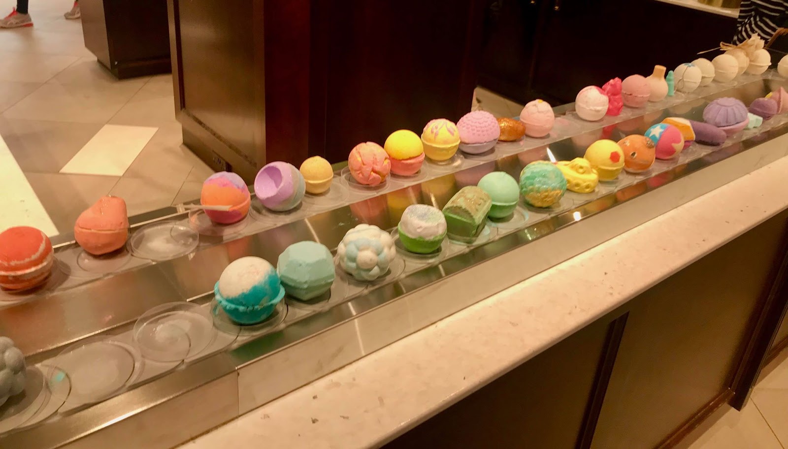 A large white rectangular shelf full of some various different coloured spherical Bath Bombs on top of some small circular white plates, on a bright background.