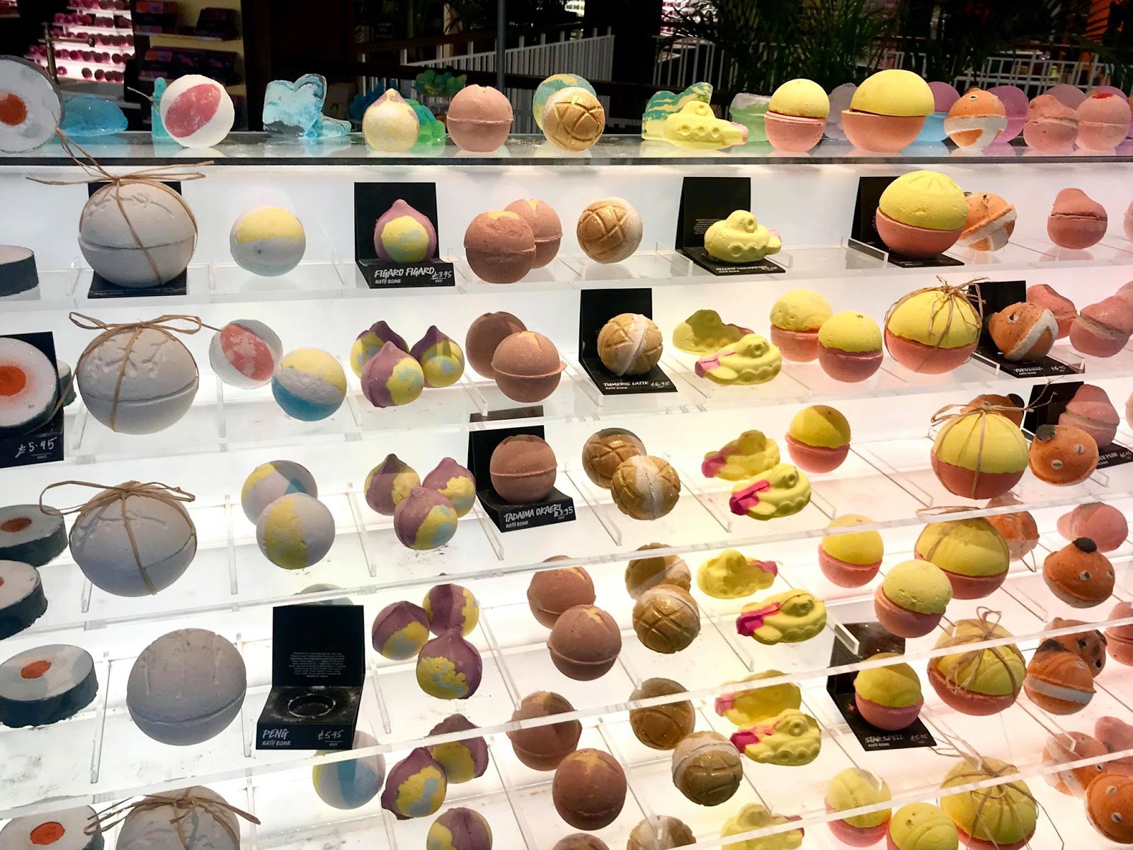 Three large rectangular clear glass shelves full of some various coloured spherical Bath Bombs, on a bright background.