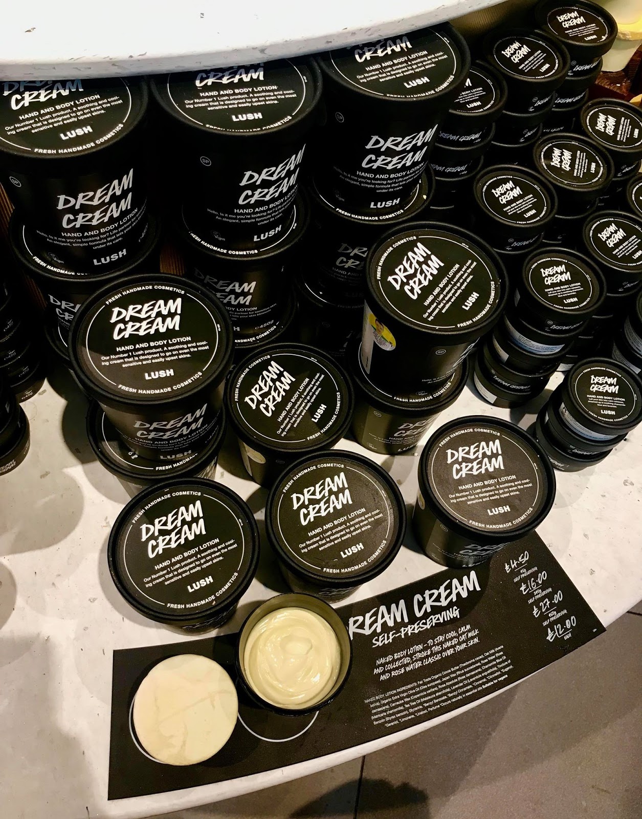 A collection of tall cylindrical black tubs that have dream cream written in bold white writing on them next to a short rectangular black board that has Dream Cream written in bold white writing on a large rectangular dark wooden shelf, on a bright background.