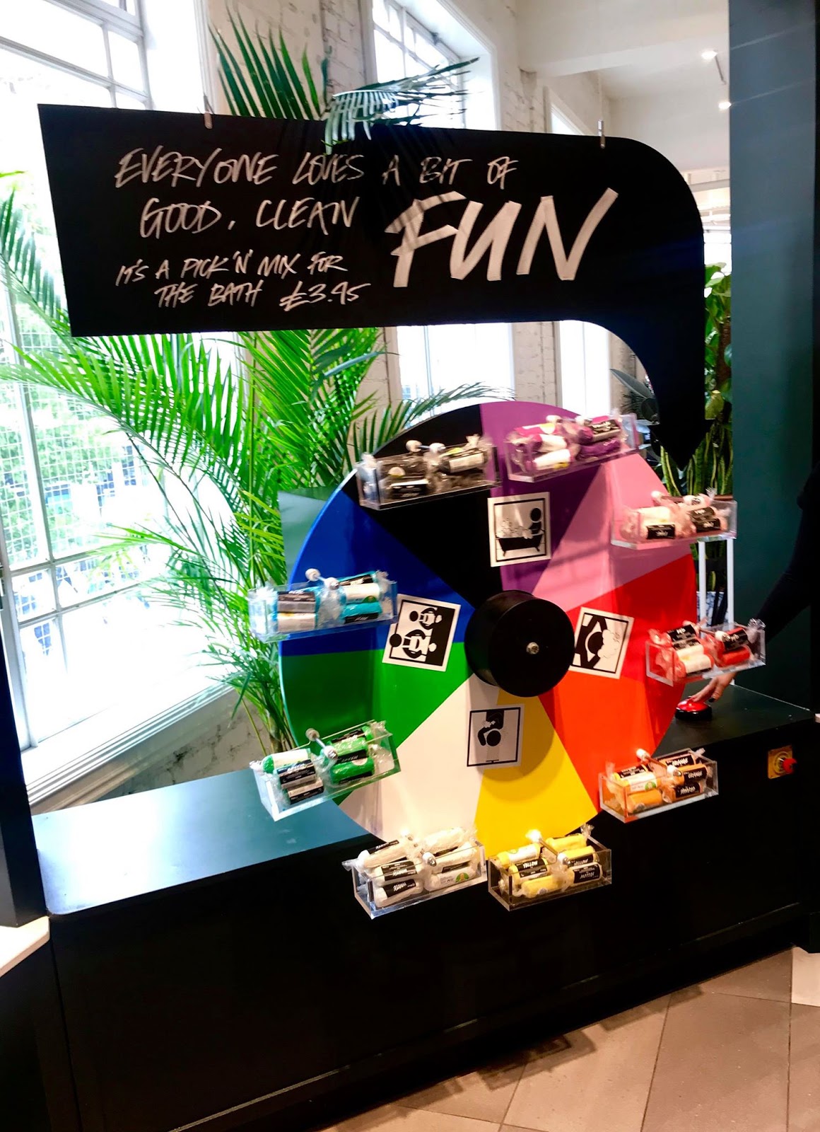 A large circular black wheel that has some little compartments around the outside of it full of some cylindrical packets of different coloured plasticine next to a large rectangular black board that has Fun written in bold white writing on it, on a bright background.