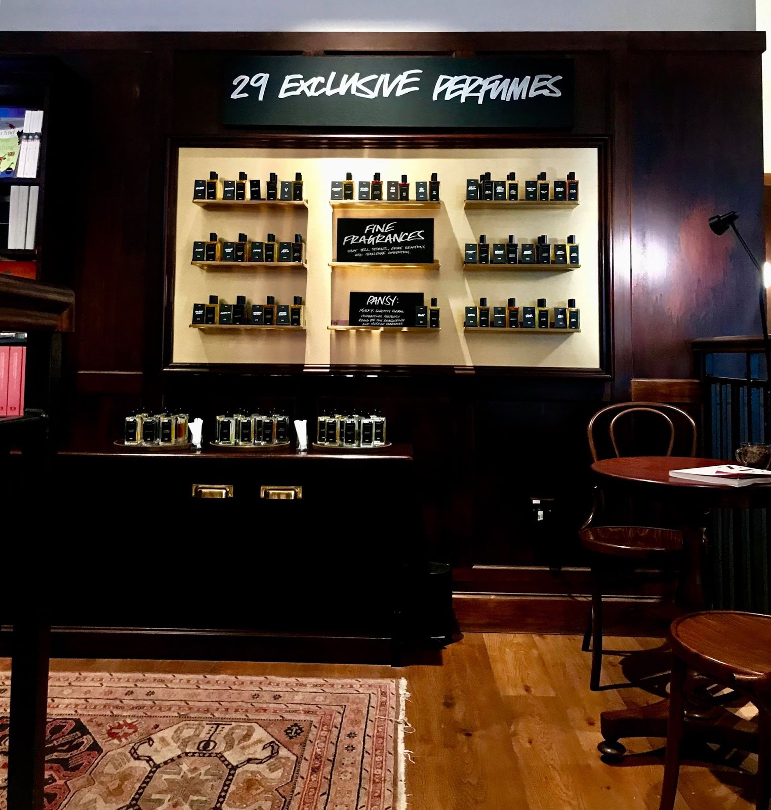A tall black wall full of some rectangular white shelves full of tall rectangular clear glass perfume bottles and a rectangular black board that has 29 new perfumes written in bold white writing on it, on a bright background.