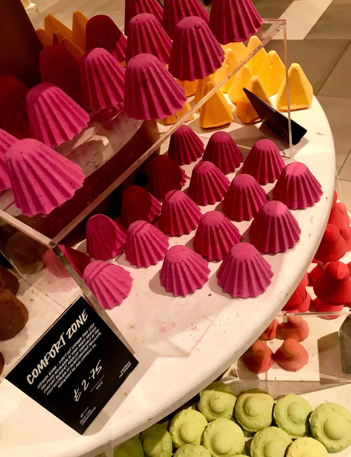 A collection of short conical bright red shower bombs on a large circular white plate next to a large rectangular black sign that has Comfort Zone Shower Bomb written in bold white writing on it, on a bright background.