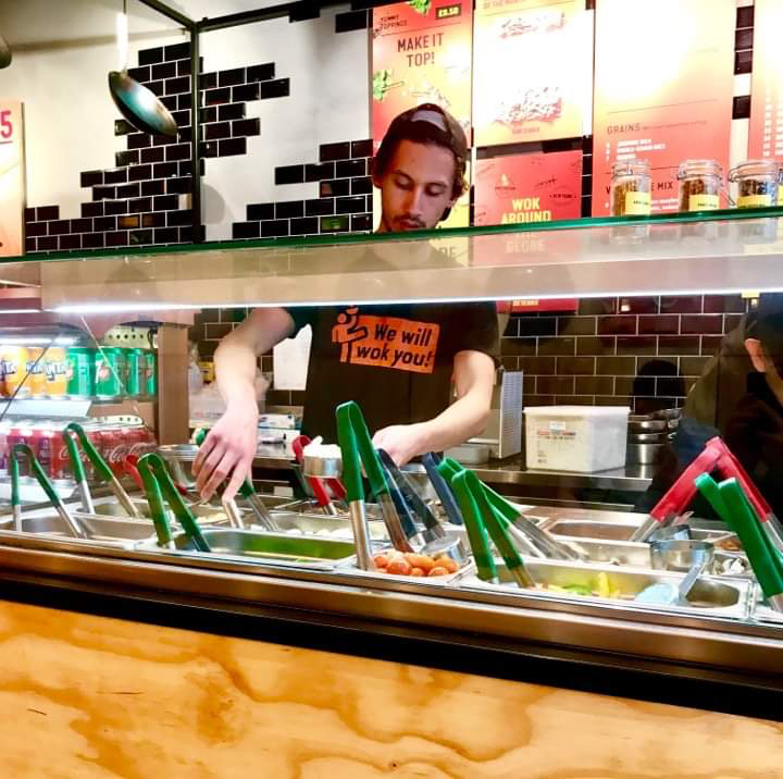 A long rectangular metal counter full of some different square compartments full of some different coloured food with a bright white light over the top of it, on a white background.