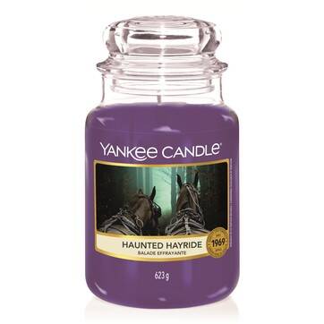 A tall glass jar full of some bright purple wax with a label that has Yankee Candle written in white writing, Haunted Hayride written in purple writing, and a picture of a field of Hay on it, on a white background.