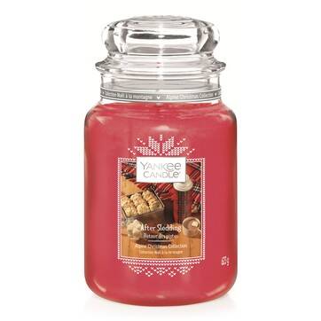 A tall glass jar full of some dark red wax with a label that has Yankee Candle written in black writing, After Sledding written in white writing, and a picture of some cookies on a Tray on it, on a white background.
