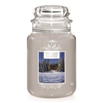 A tall glass jar full of some ice blue wax with a label that has Yankee Candle written in black writing, Candlelit Cabin written in white writing, and a picture of a large brown log cabin on it, on a white background.