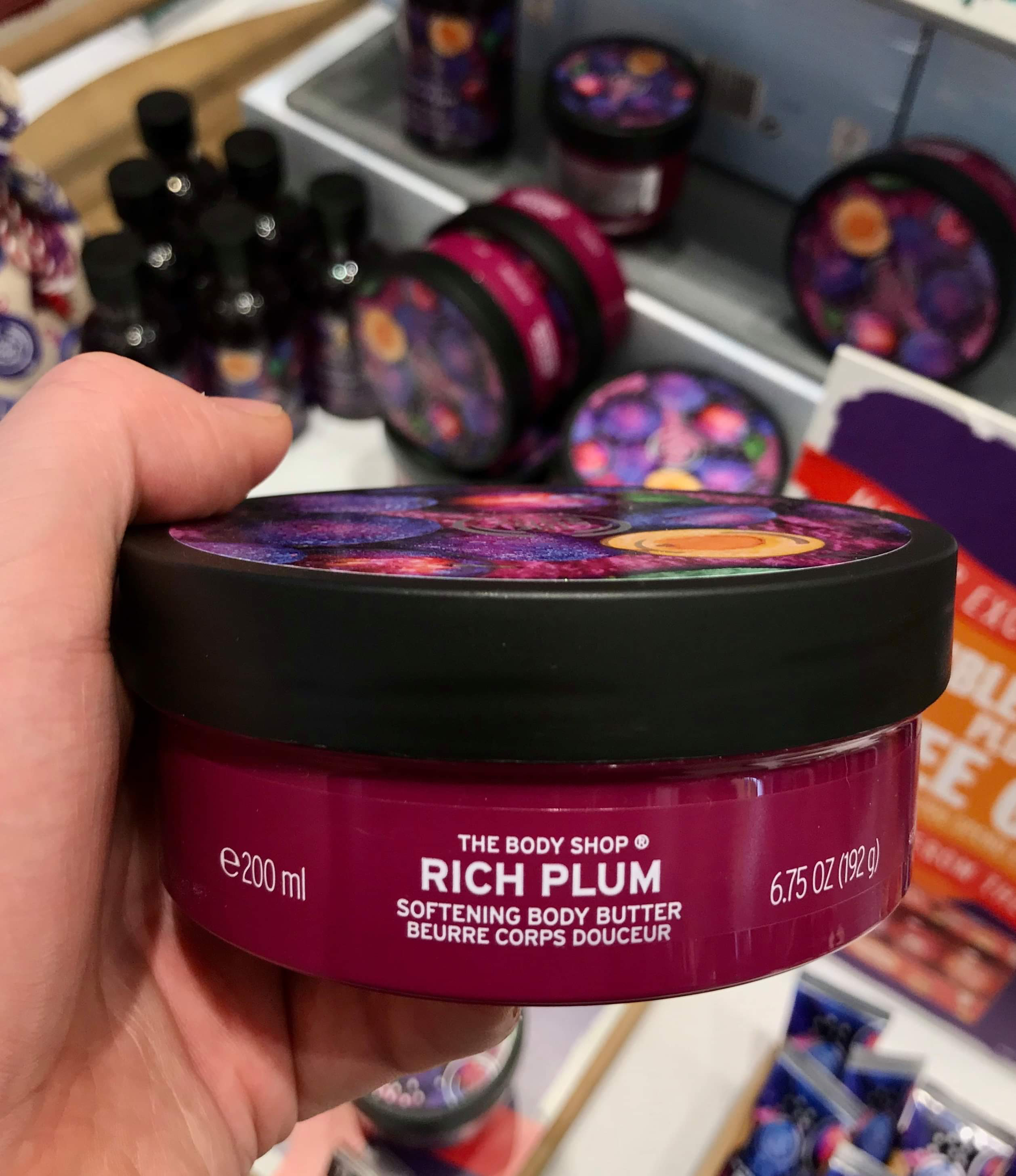 A large circular dark purple plastic Tub with a white plastic lid and a bright red label that has the body shop rich plum body butter written in bold white writing and some pictures of some plums on it, on a white background.