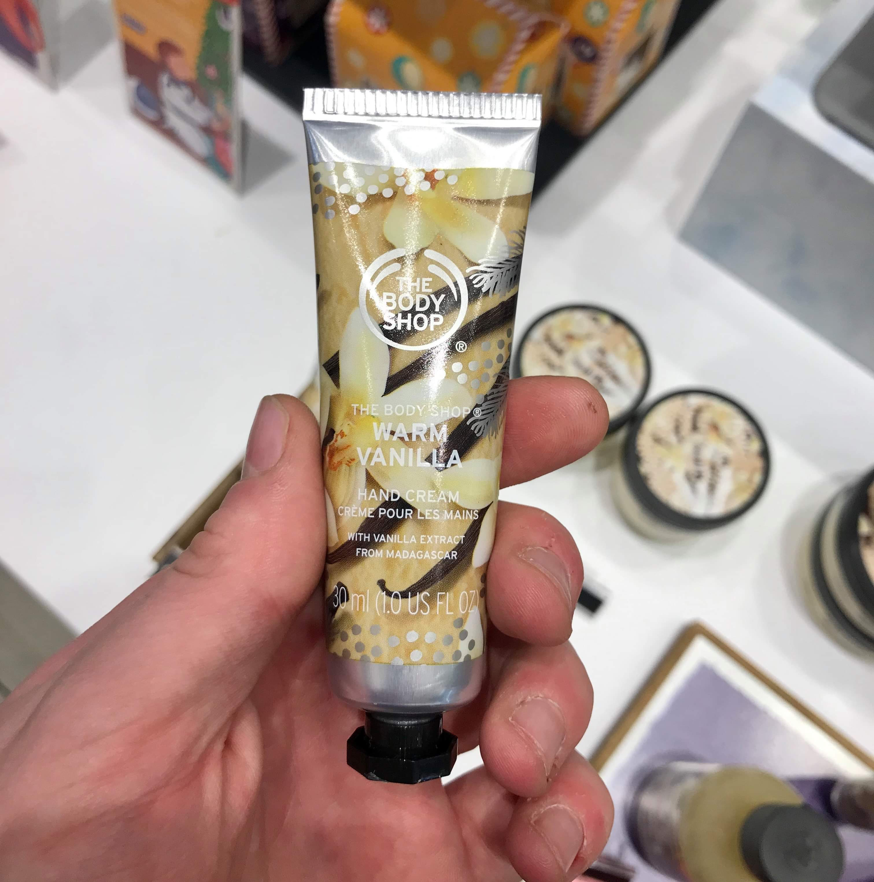 A small thin conical silver plastic tube with a black lid that has the body shop warm vanilla hand cream written in bold white writing and some pictures of some vanilla pods on it, on a white background.