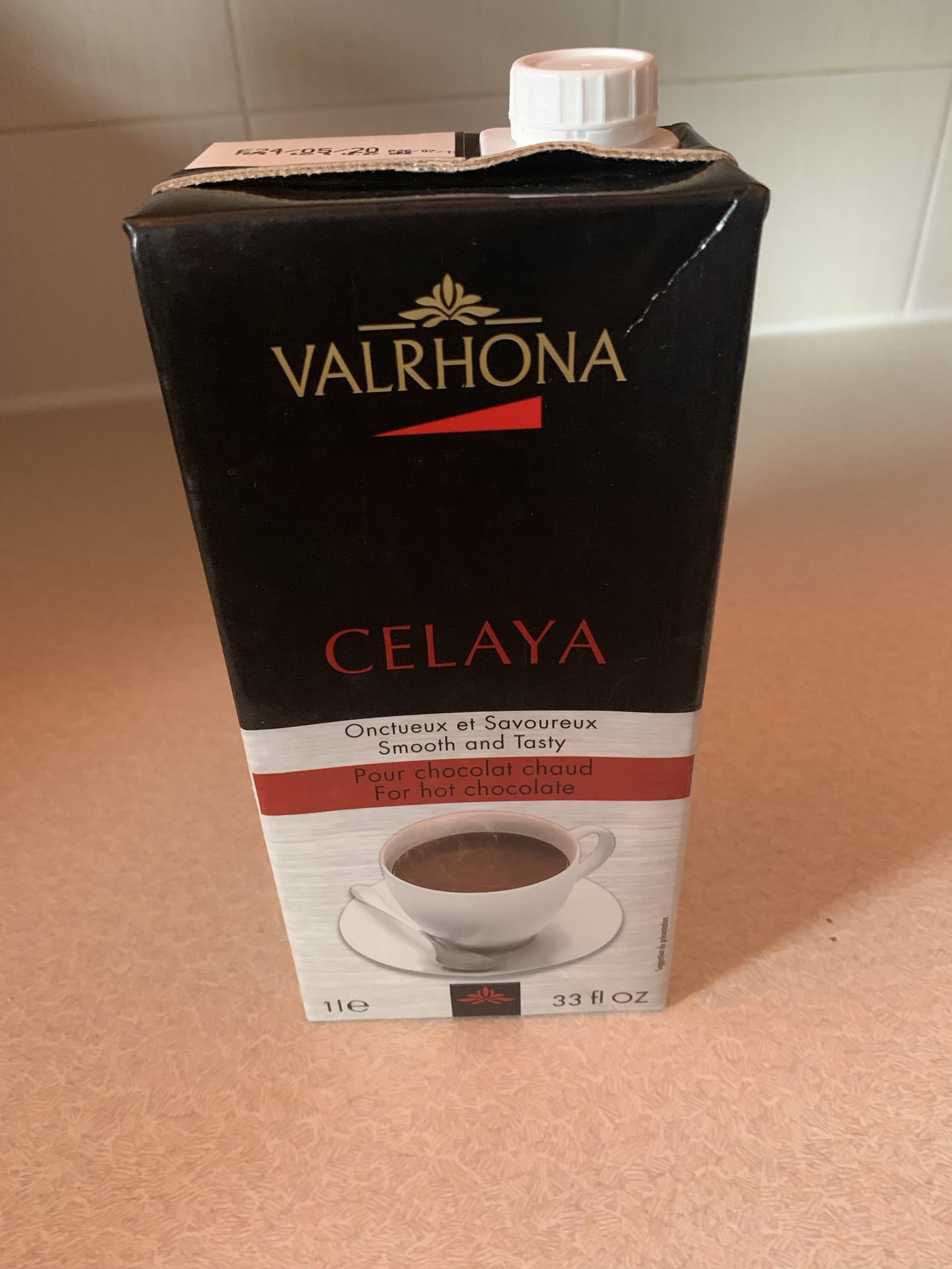 A tall rectangular black and white cardboard carton that has Valrhona Celaya Liquid Hot Chocolate written in gold and red writing on it, on a white background.