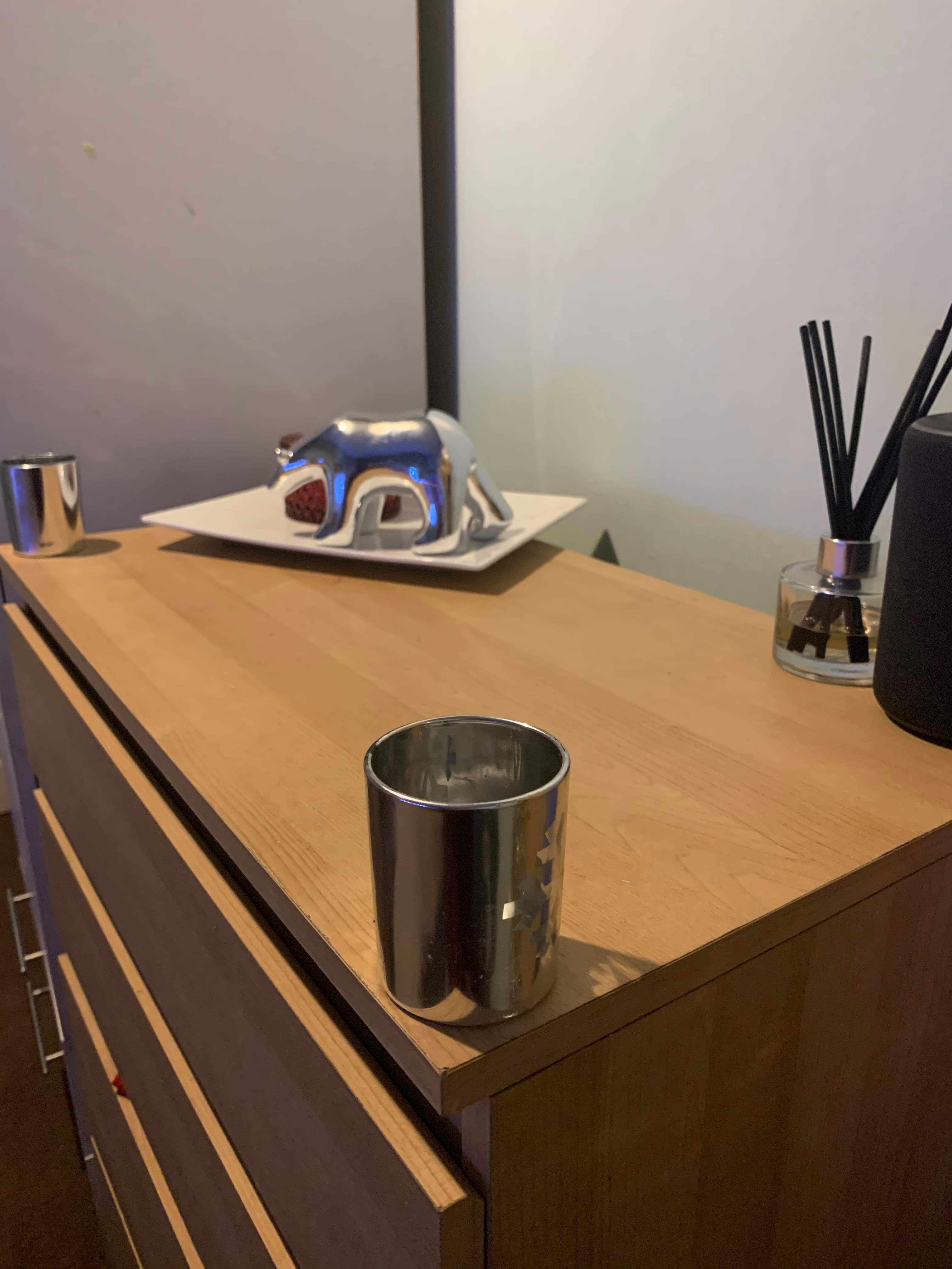 A large silver Polar Bear ornament stood on all fours next to a short cylindrical cut glass bowl on a large rectangular white plate next to two short cylindrical glass votive candle holders that have snowflakes all over them and a tea light inside them on a light wooden chest of drawers, on a white background.