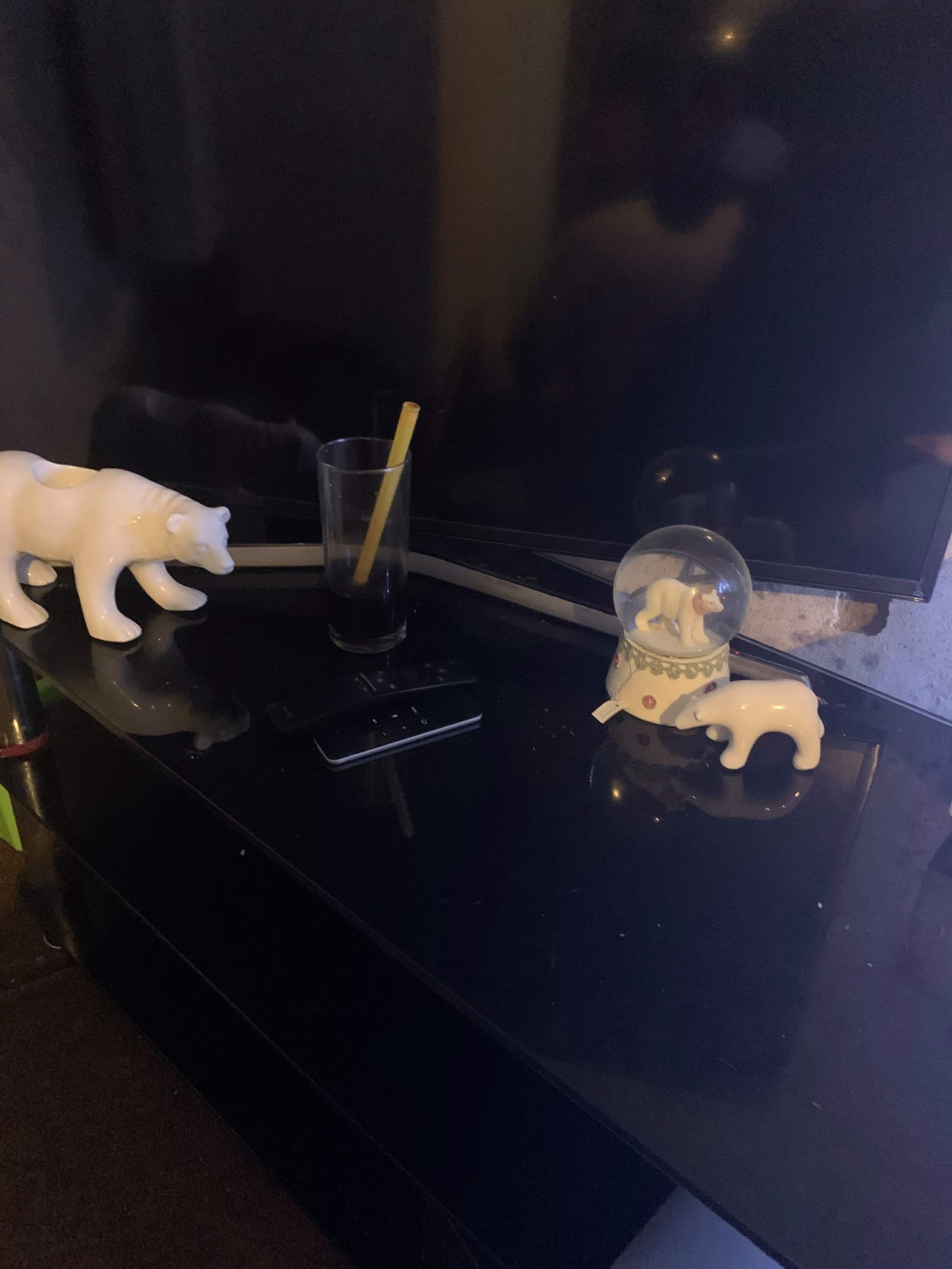 A large bright white snow globe that has a Polar Bear stood on all fours inside it next to a small white ceramic polar Bear ornament also stood on all fours and a large white ceramic Polar Bear Tea Light holder stood on all fours in front of a large black TV on a black Glass Cabinet, on a white background.
