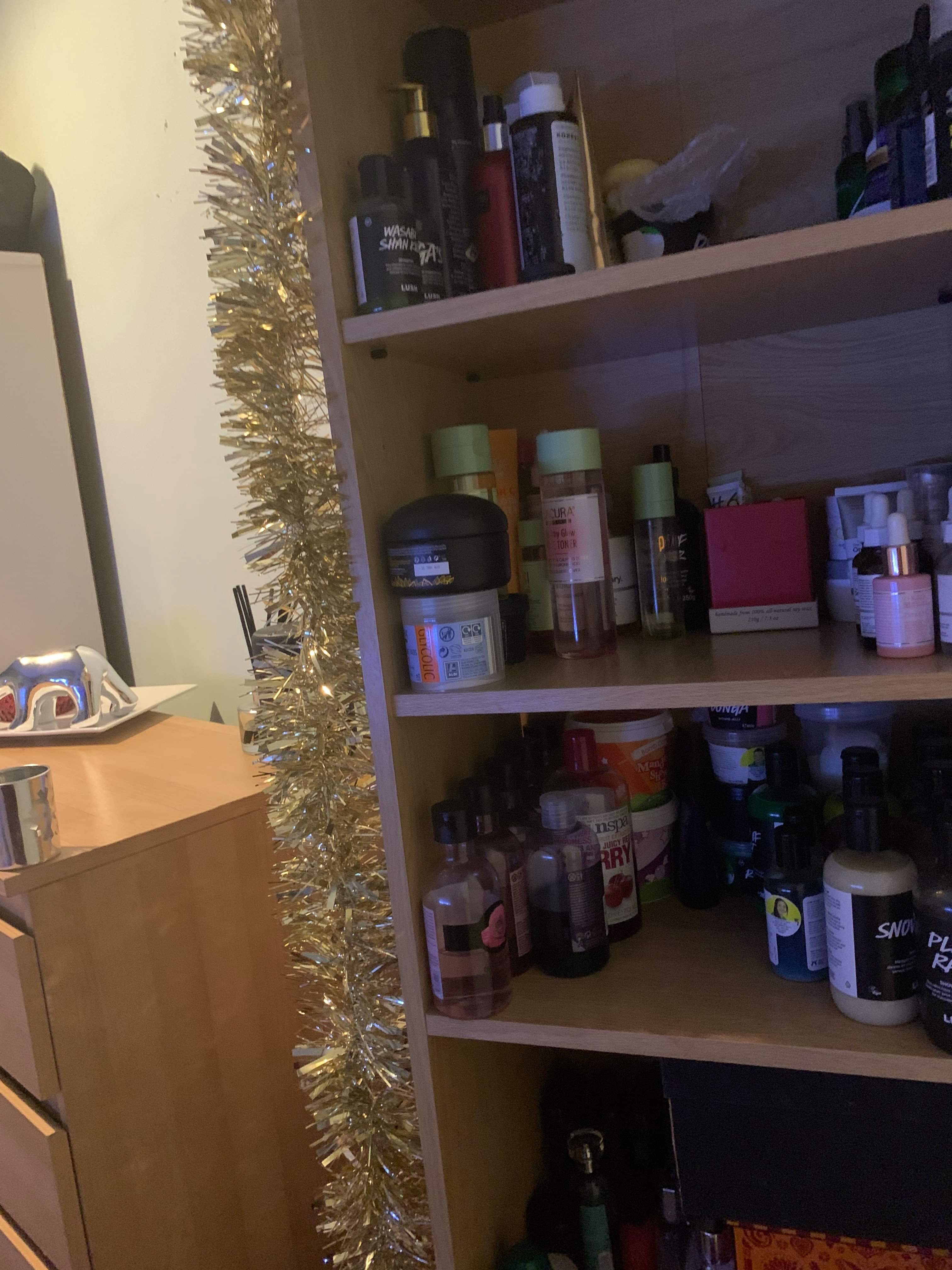 Some shiny silver tinsel wrapped around a tall light wooden rectangular bookcase full of some various different shaped bottles, on a light background.