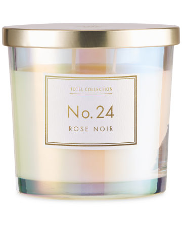 A medium cylindrical clear glass jar full of some bright white wax with a shiny gold metal lid and a white label that has Aldi Hotel Collection Rose Noir Candle written in bold gold writing on it, on a white background.