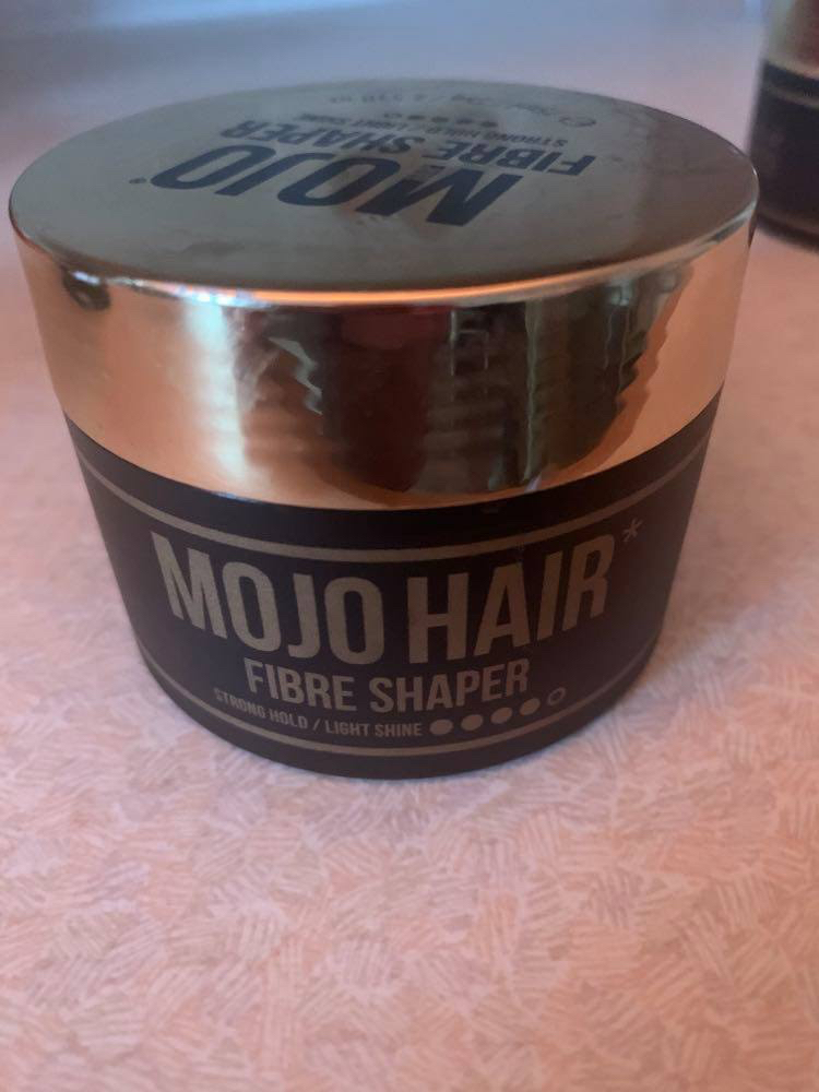 A short cylindrical matte black plastic tub with a glossy gold plastic lid that has Mojo Hair* Fibre Shaper written in bold gold writing on it, on a white background.