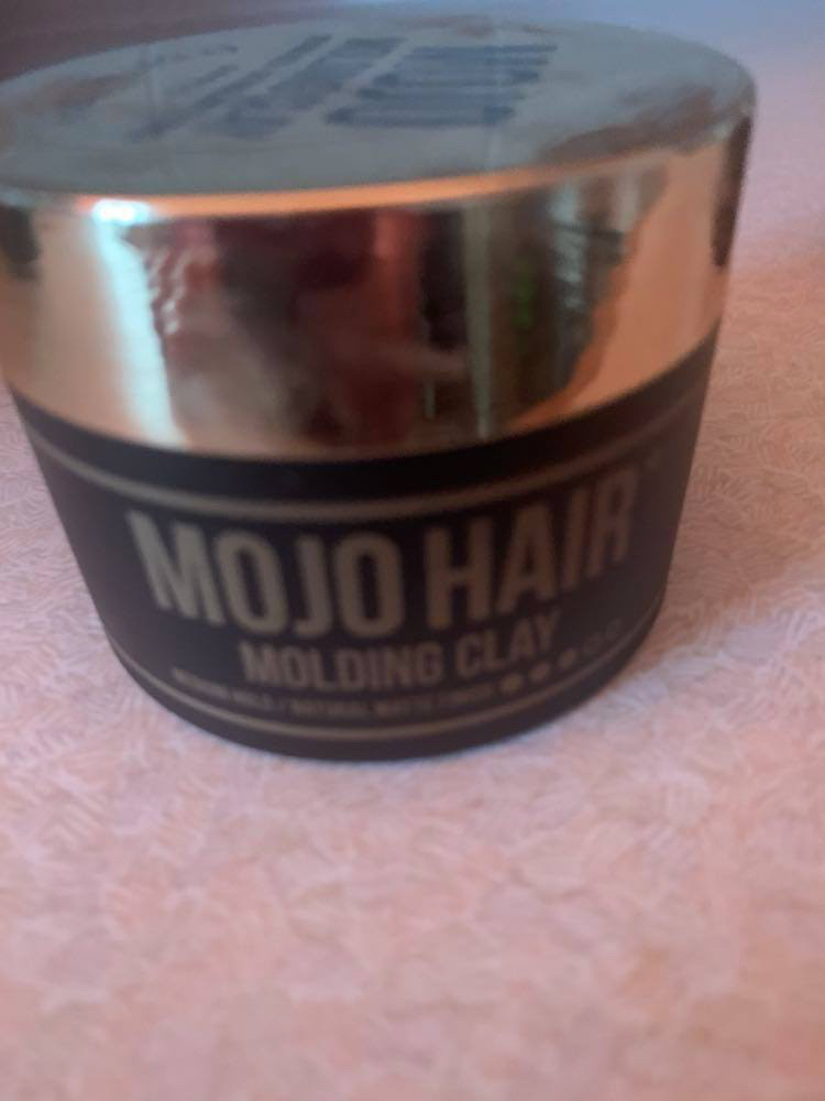 A short cylindrical matte black plastic tub with a glossy gold plastic lid that has Mojo Hair* Moulding Clay written in bold gold writing on it, on a white background.