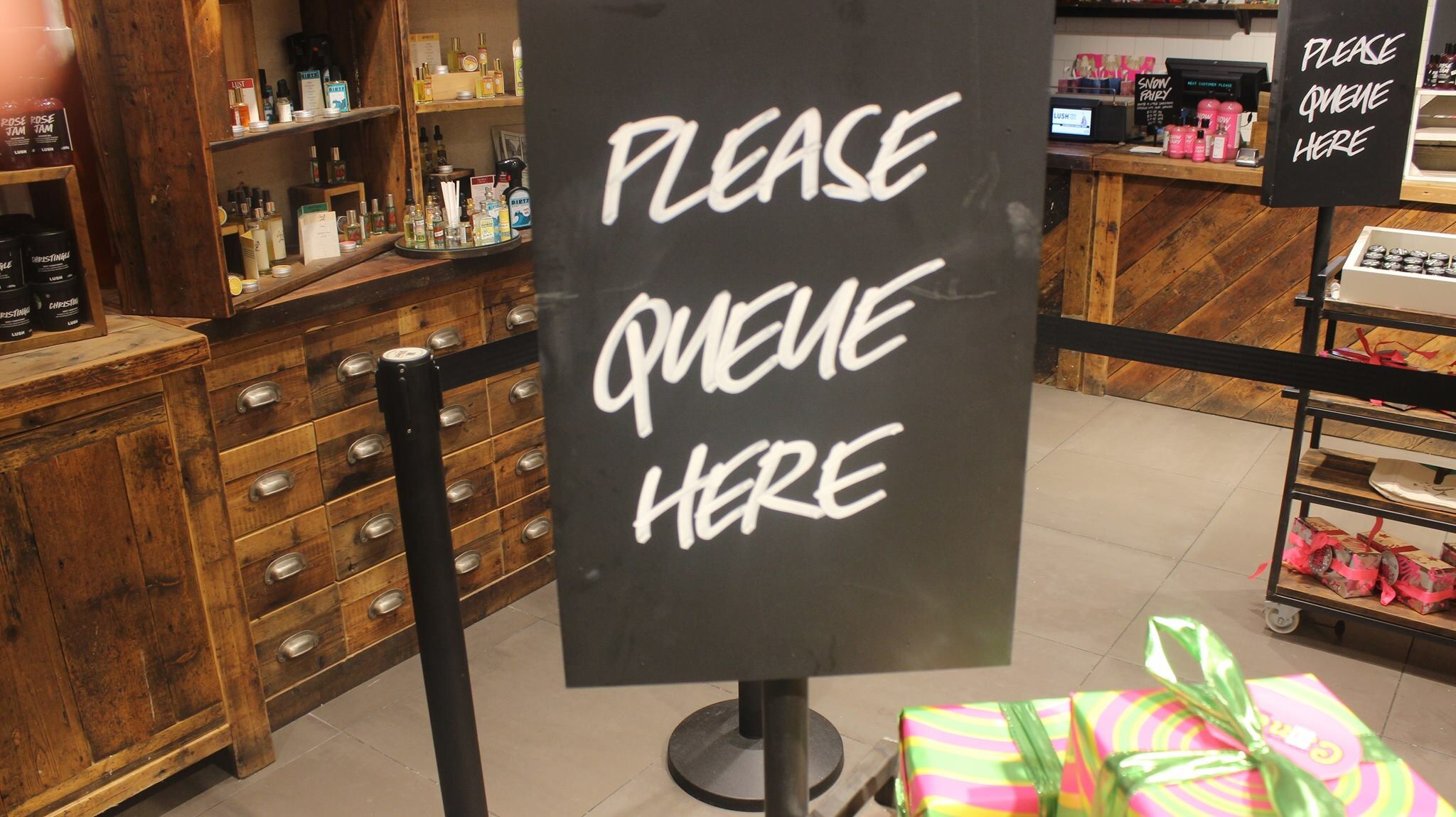 A large rectangular black sign that has Please Queue Here written in bold white writing on it attached to some Black Rope, on a white background.