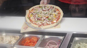 A person holding a Pizza in front of some sliced vegetables.