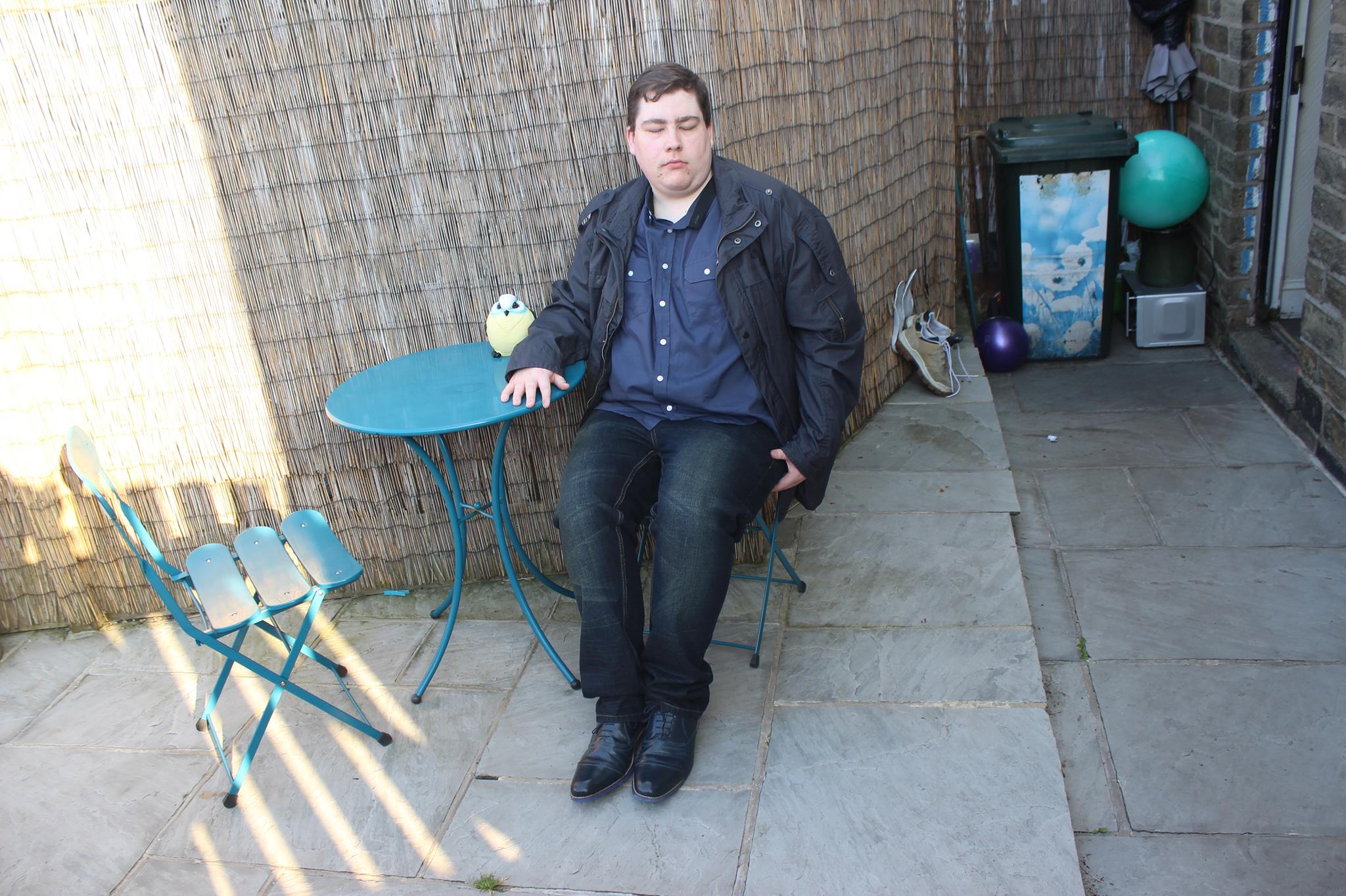 A person sat in a garden chair wearing a jacket, a short sleeved shirt, a pair of jeans, and some shoes.