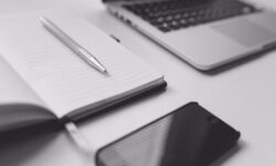 A black iPhone, a silver pen and a notebook next to a laptop on a white rectangular table, on a white background.