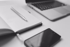 A black iPhone, a silver pen and a notebook next to a laptop on a white rectangular table, on a white background.
