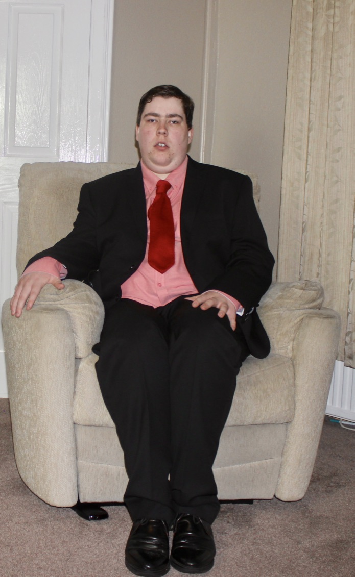 Outfit Of The Day | Time To Suit Up A full body picture of myself, wearing a black suit, sat on a grey chair, with a grey and white background.