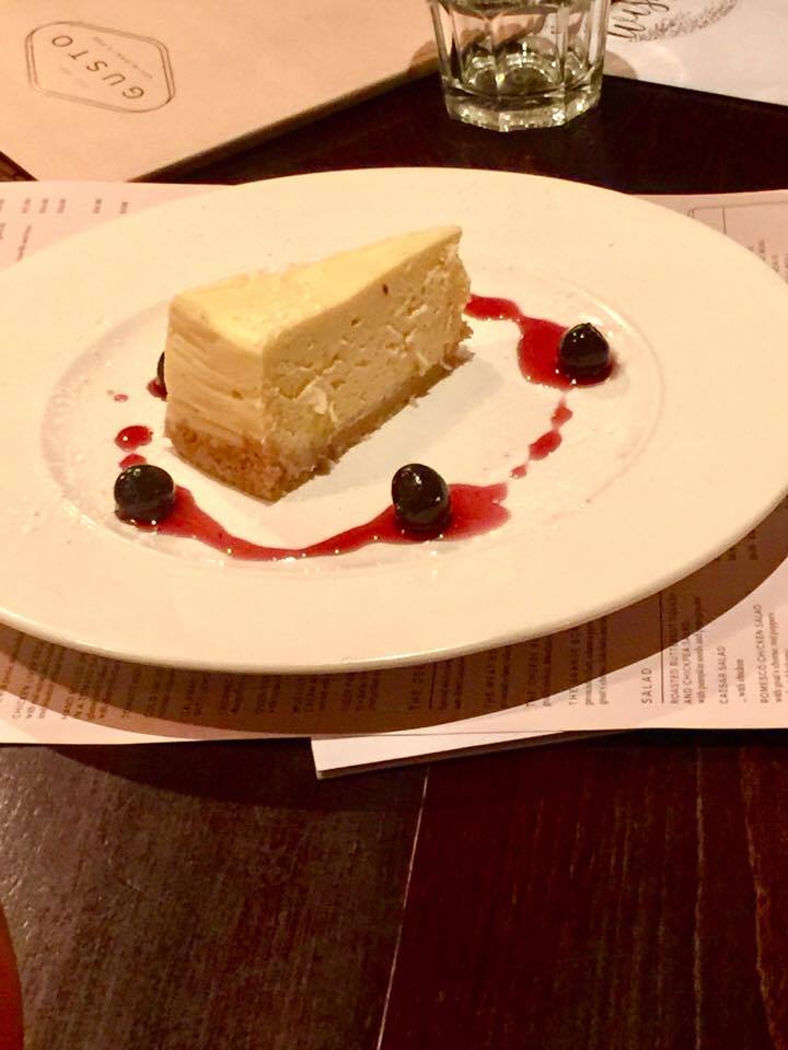 Put Some Gusto Into It | Gusto Cookridge Autumn Winter 2016 Menu Preview A creamy white wedge of cheesecake, with a tan base, and some red sauce circling the plate, with some red cherries, on a white plate, on a brown and white background.