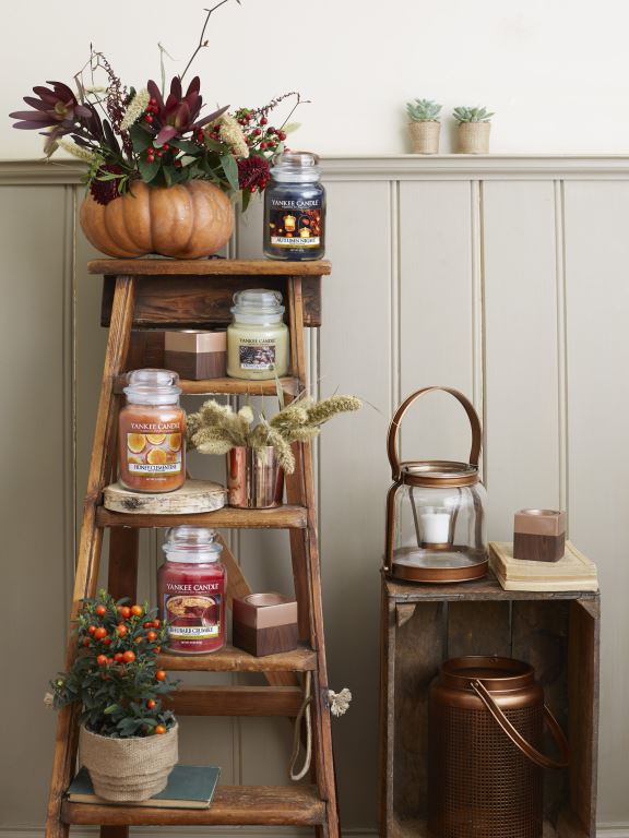 A collection of candles in glass jars, on wooden shelves, and some golden tea light holders, on a white background.
