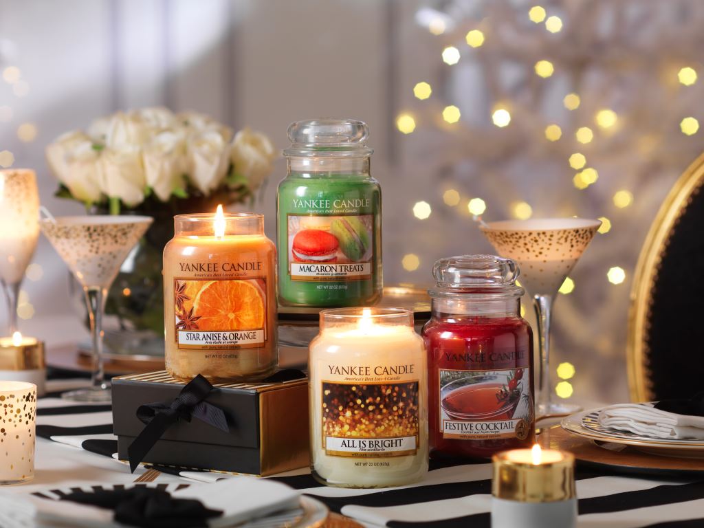 The Best Smelling Party Around | Yankee Candle Holiday Party Collection A collection of four tall glass jar candles that include Yankee Candle All Is Bright, Yankee Candle Star Anise & Orange, Yankee Candle festive Cocktail, and Yankee Candle Macaron Treats, on a wooden table, with some fairy lights in the background.