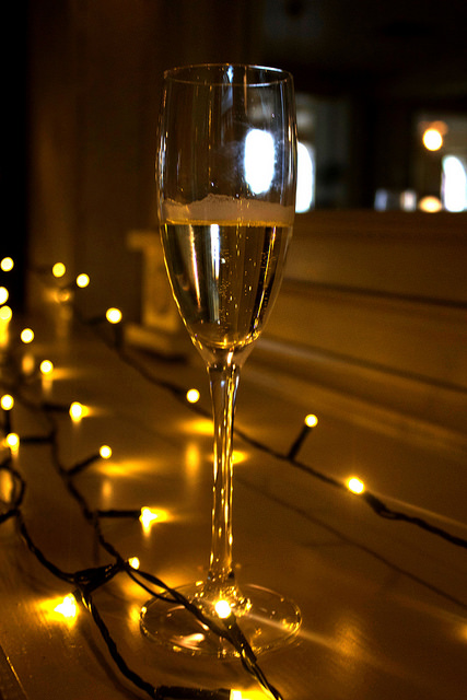 #BlindGuyAdventures | Fashion, Nails, and Prosecco. A clear long stemmed glass of clear bubbly Prosecco, on a wooden background, with some fairy lights in the background.