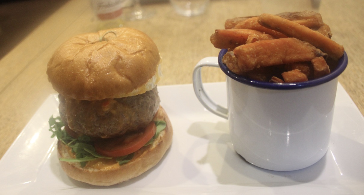 A burger with dark brown meat with some green lettuce leaves and a red Tomato, with some golden sweet potato fries in a white mug, on a white plate, on a dark tan background.