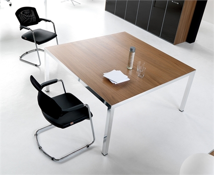 A light brown coloured desk, with a bottle and some paper on top of it, opposite a brown bookcase, and another black leather chair, on a white background.