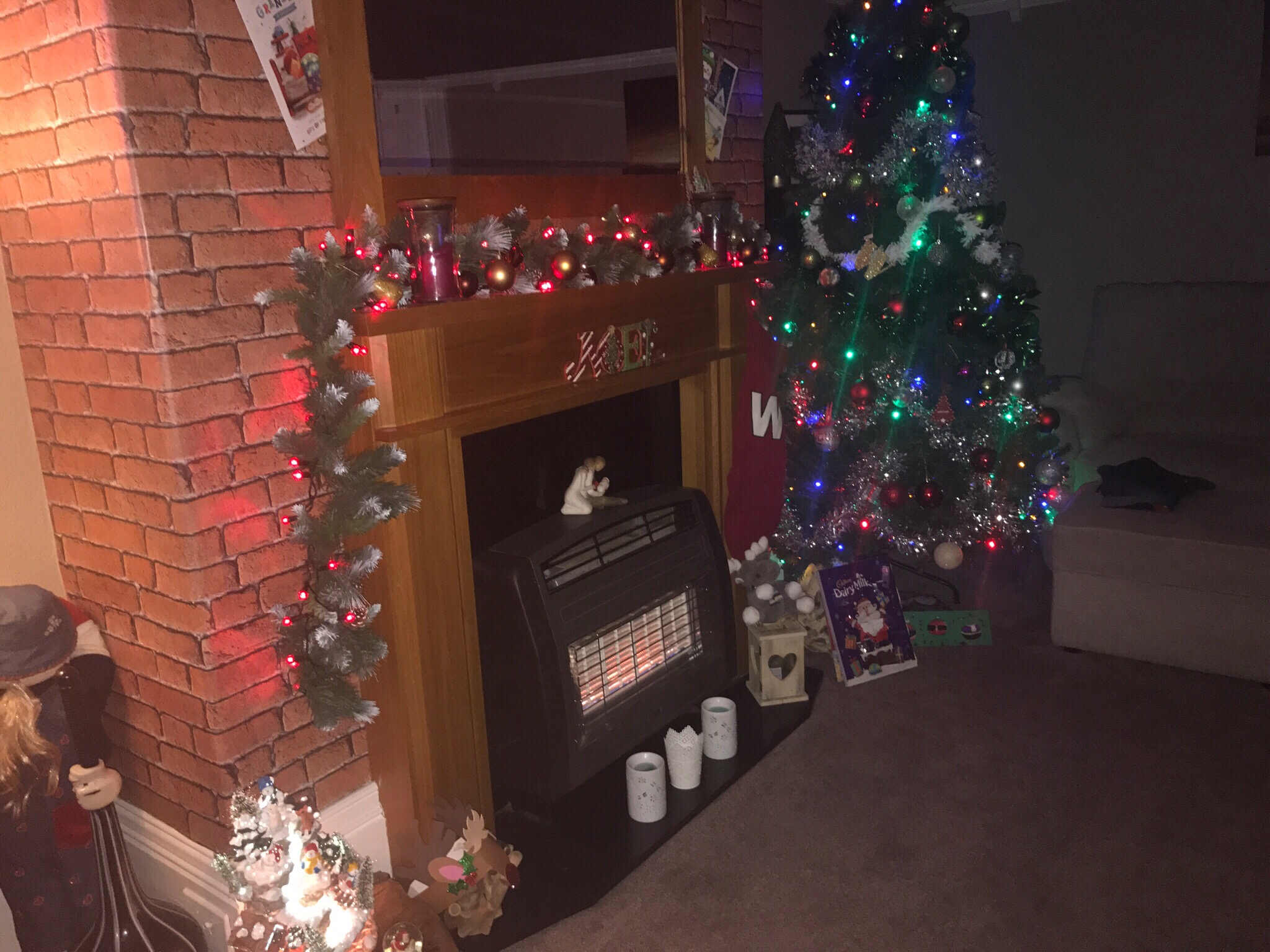 A dark green Christmas tree covered in some blue lights next to a dark brown fireplace and a light brown mantle piece covered in some red lights, on a white background.