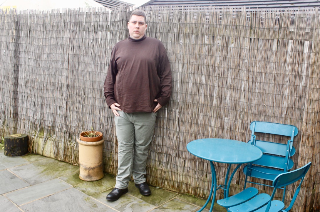 A person wearing a chocolate brown roll neck top , a pair of green stretched straight chinos, a black leather hook bracelet, and a pair of black leather boots, on a wood background with wood plants and a blue table and chairs in the background.