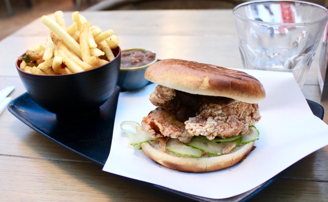 An Asian Influenced World | Bar Soba Leeds Spring 2017 Menu A tan burger bun filled with a golden breadcrumbed Chicken patty, with hints of green and white Cucumber next to a tall cylindrical black pot full of golden thin fries and a wide shallow circular pot of dark gold Katsu sauce on a white rectangular plate, on a dark background.