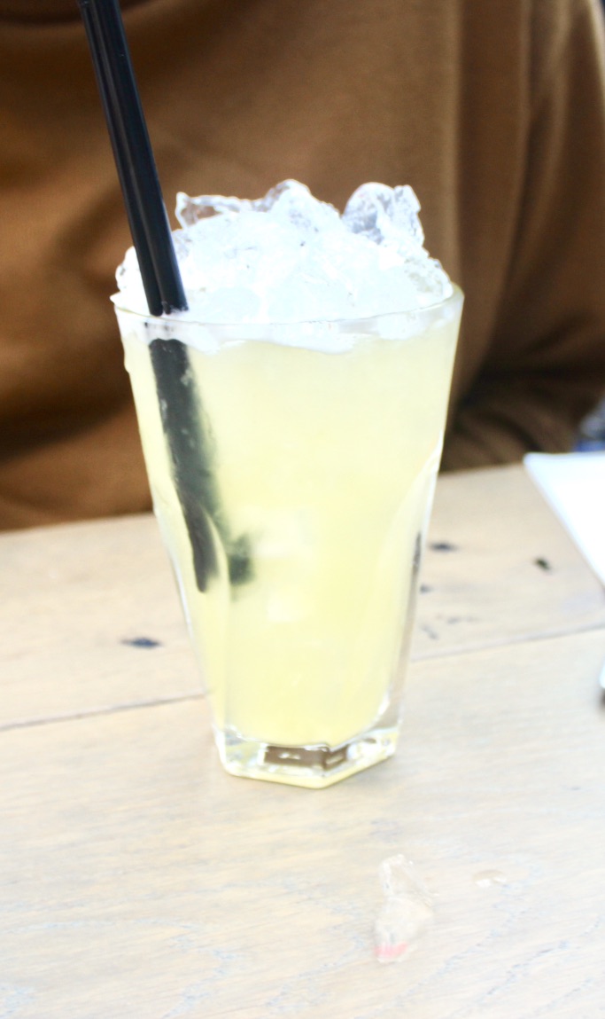A tall glass full of pale yellow and green liquid with some clear shards of ice and two black straws on a tan table, on a dark background.