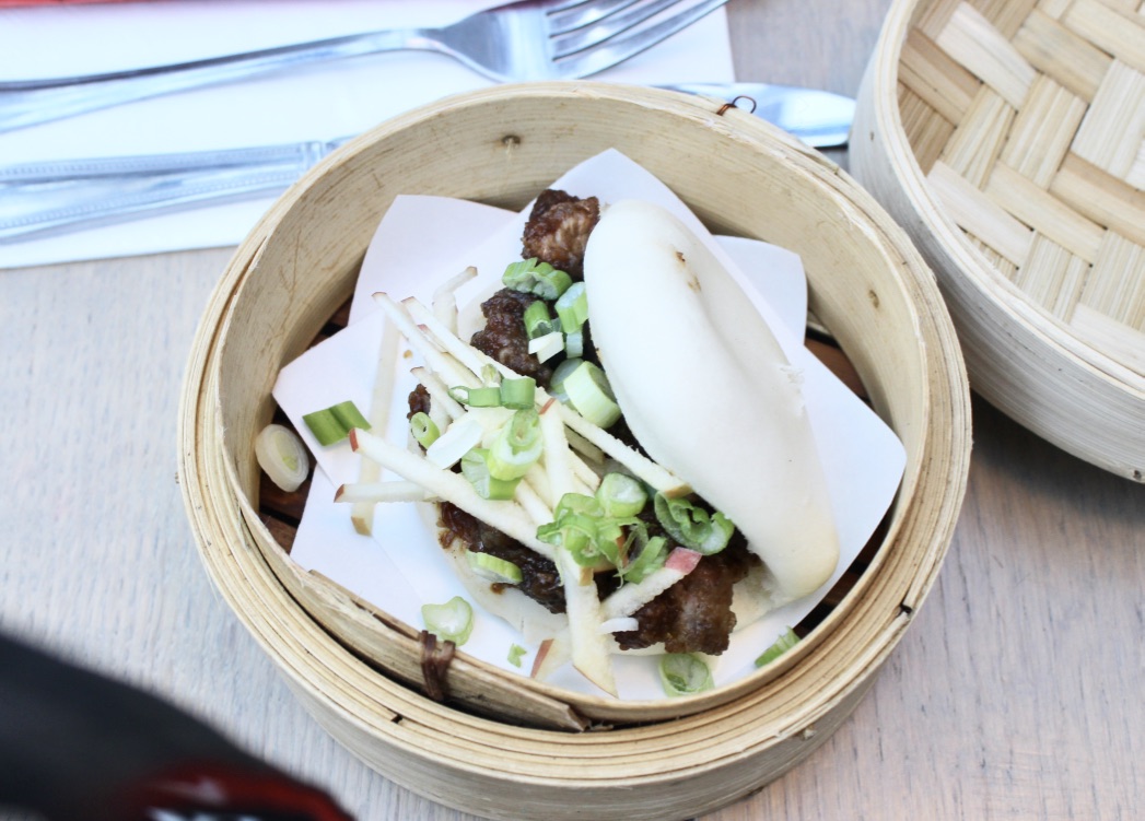 Sweet, Sugar, Candy Man! | Smith & Sinclair Leeds Pop Up Store Launch with The Alchemist Trinity Leeds A pale white bun filled with dark brown Pork, white strips of Apple, and green and white circles of Spring Onion in a tan bamboo basket, on a white and blue background.