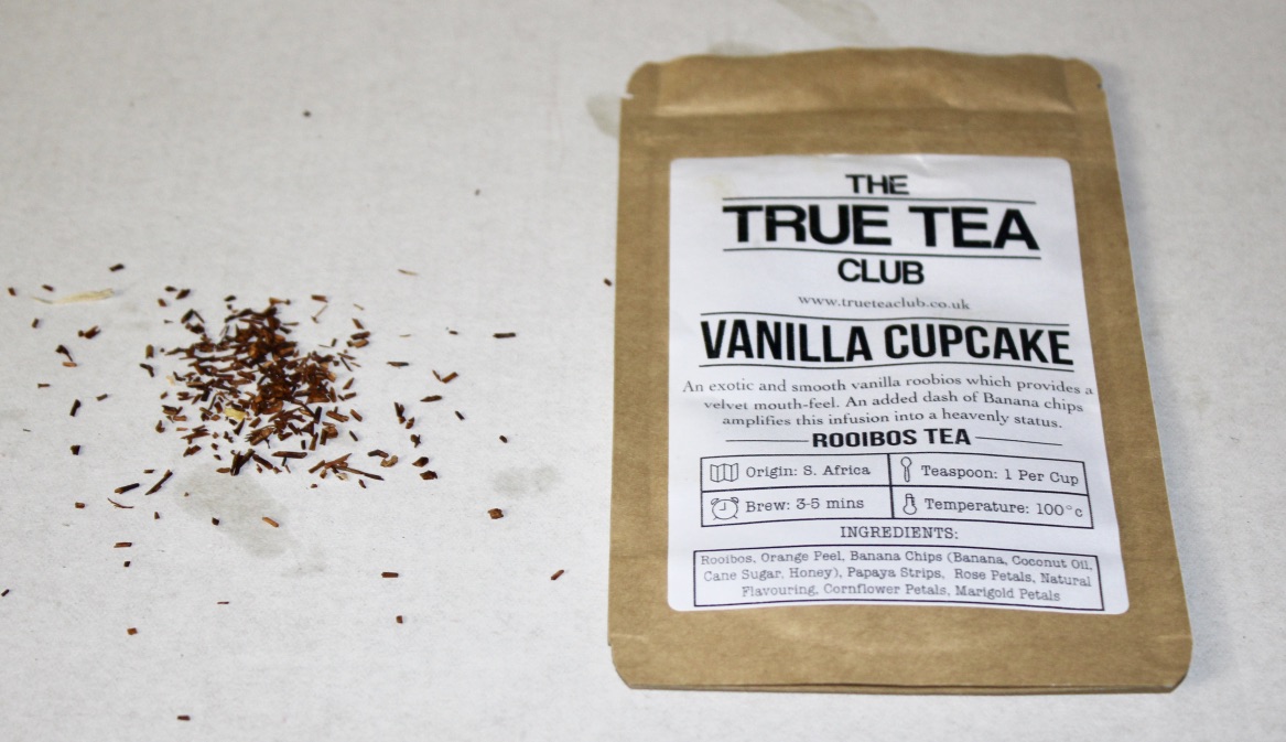 Tea Tastes Terrific | Whittard of Chelsea Apple Crumble Loose Leafed Tea A rectangular brown pouch with a white label that has True Tea Club Vanilla Cupcake Rooibos Loose Leafed Tea written in black writing on it, with some black Tea Leaves next to the pouch, on a white background.